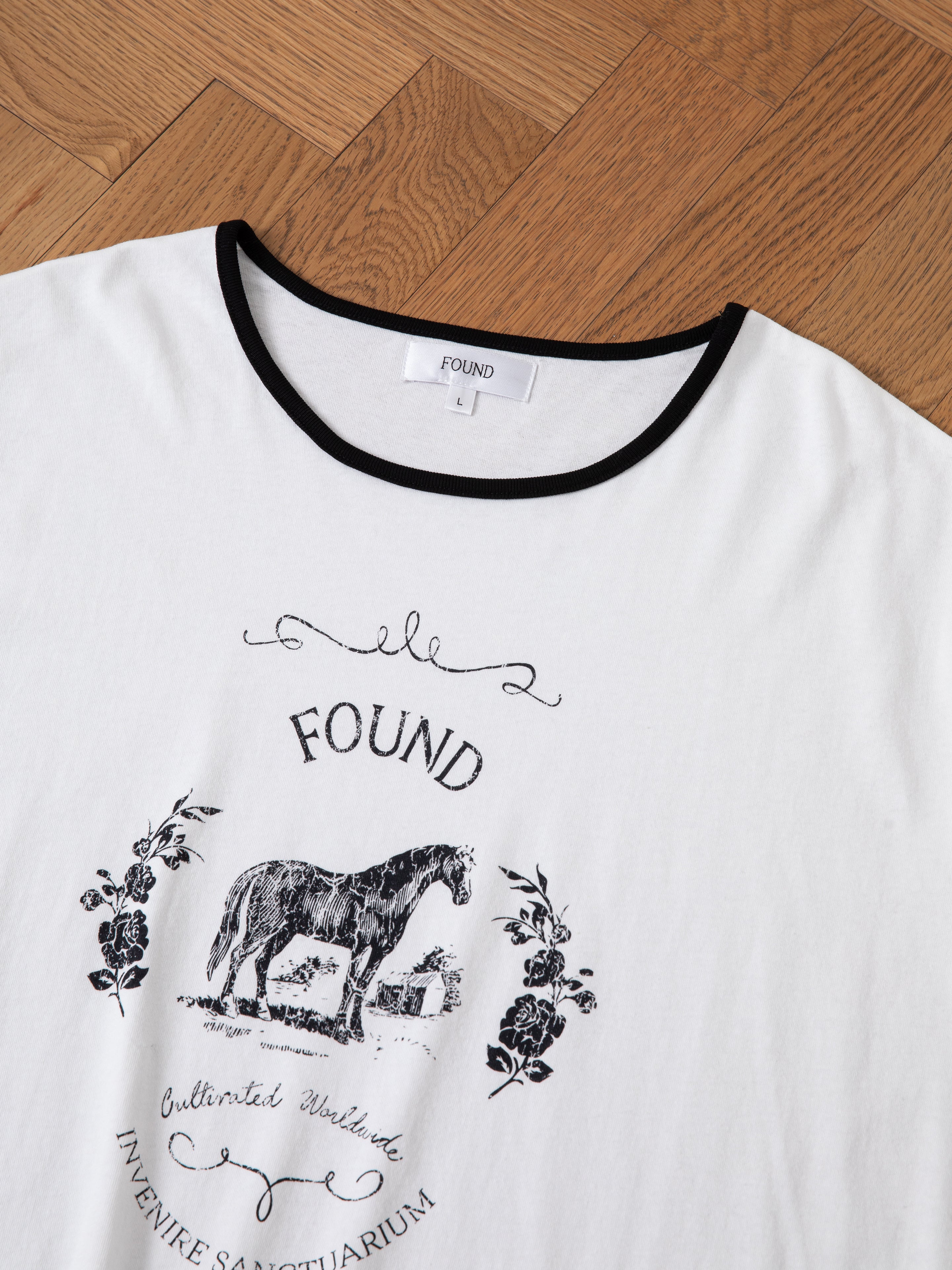 White 100% cotton Horse Crest Ringer Tee by FOUND, featuring a black collar trim and a distressed screen print of a horse graphic alongside decorative script and floral elements. The shirt is laid flat on a wooden floor.