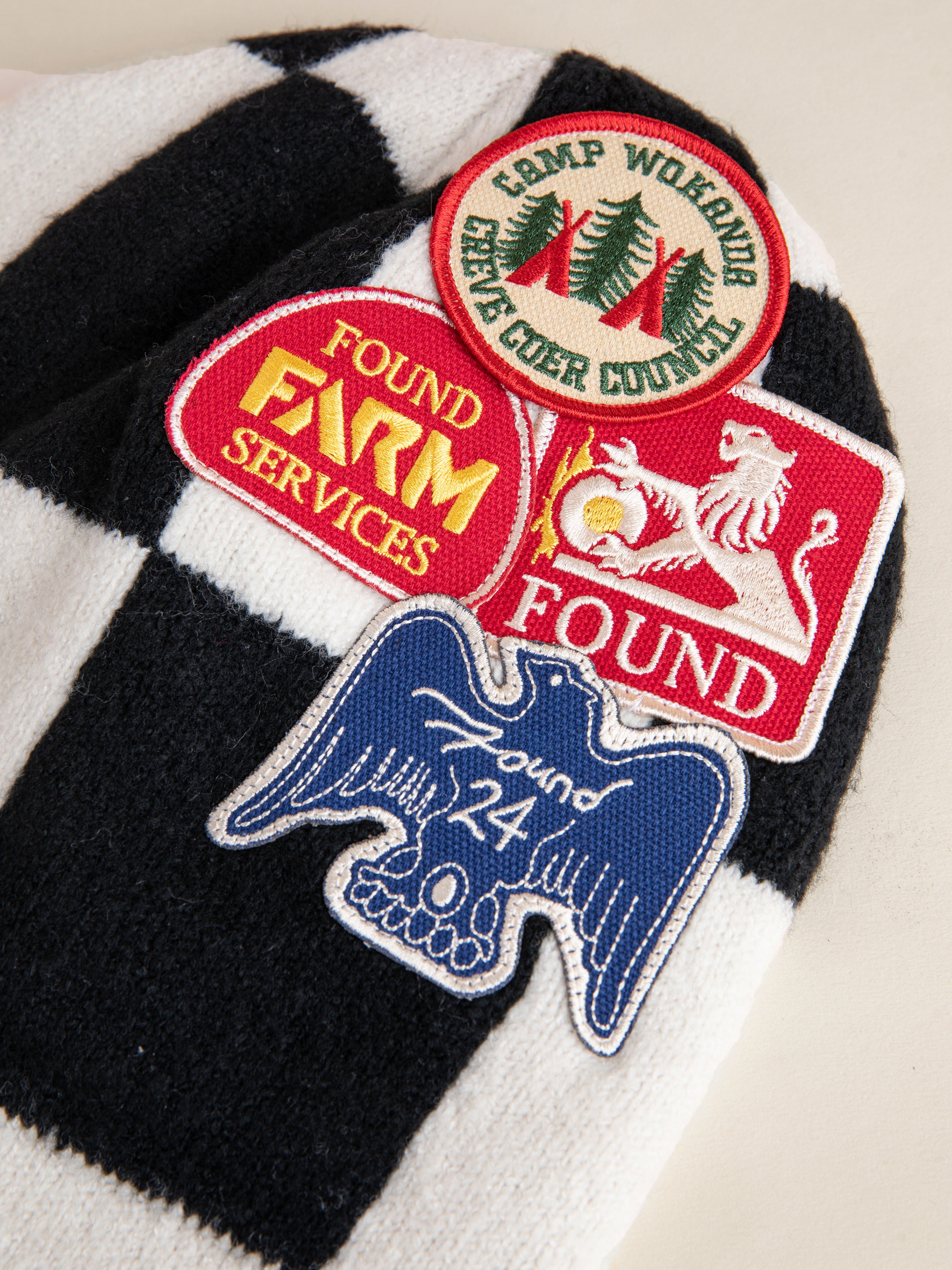 A striking black and white checkered fabric embellished with four embroidered patches: a red patch depicting a lion, another featuring trees, one inscribed with "Farm Services," and a blue patch highlighting an eagle alongside "Found 24." Ideal for making your FOUND Check Multi Patch Beanie truly stand out.