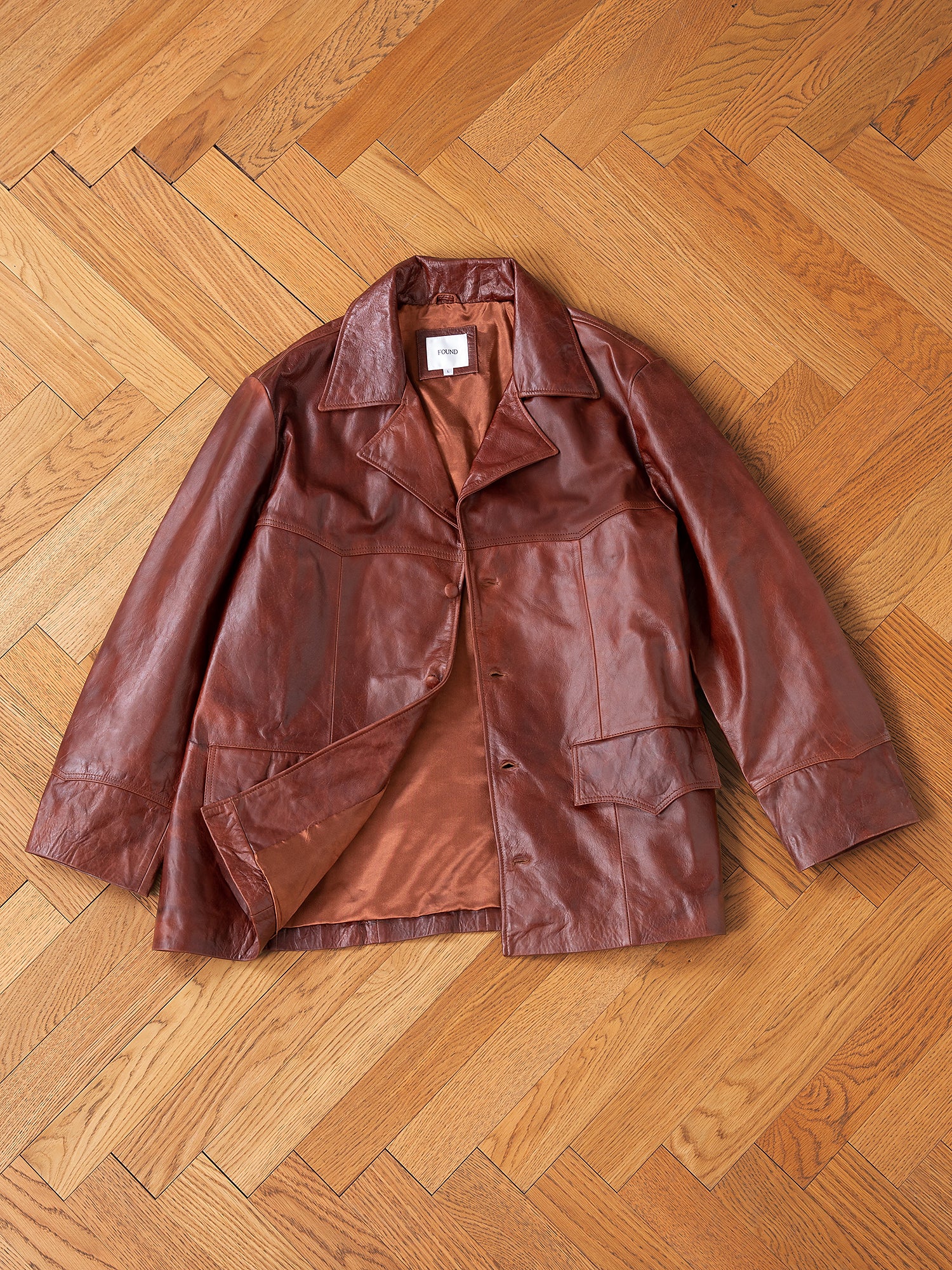 A unisex brown leather jacket with a collar and two front pockets on a wooden parquet floor embodies the FOUND Claret Western Oversized Leather Blazer.