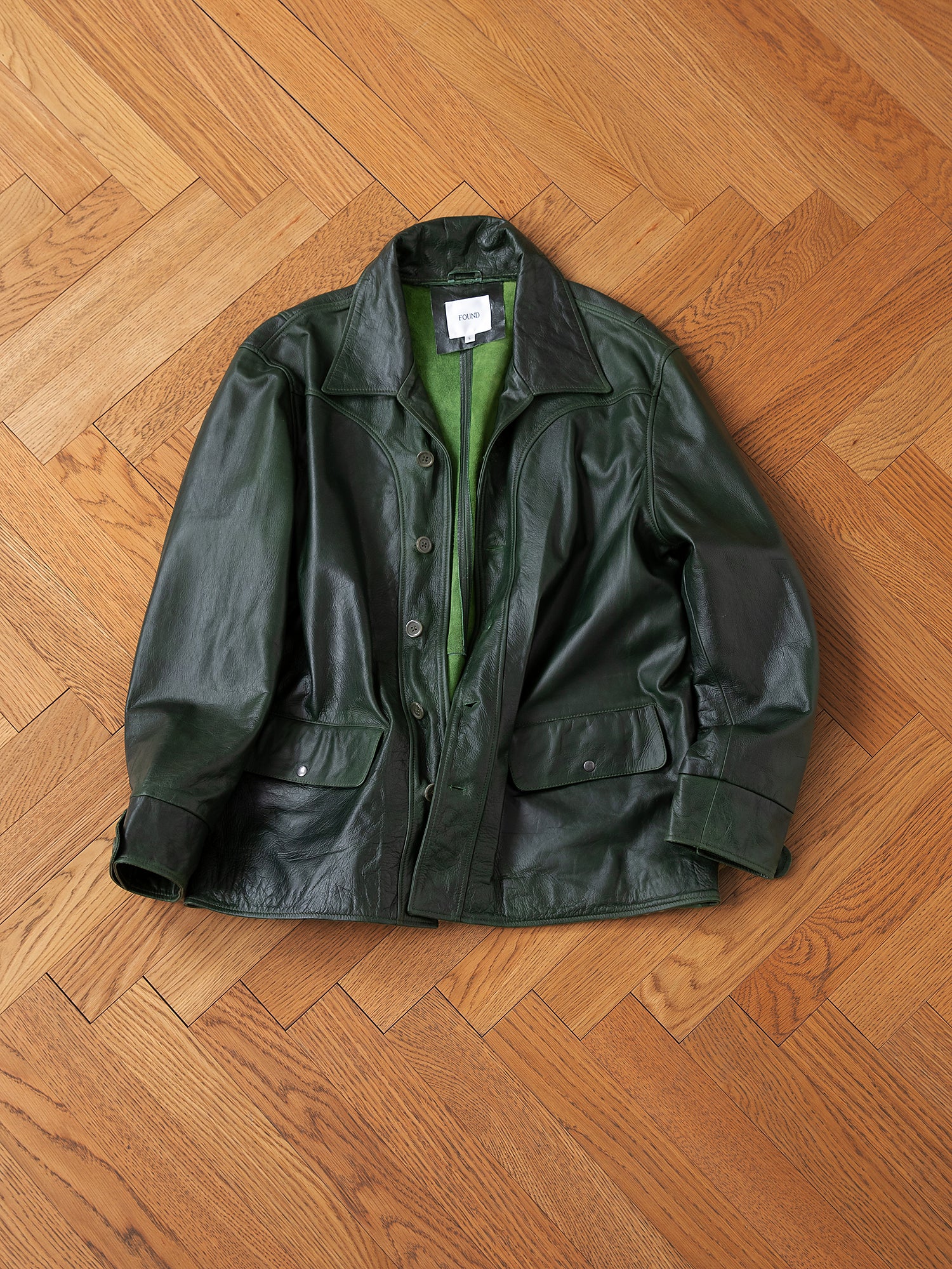 The FOUND Leather Field Jacket in emerald green, made from hand-distressed, ethically sourced leather with front pockets, is displayed flat on a herringbone wooden floor.