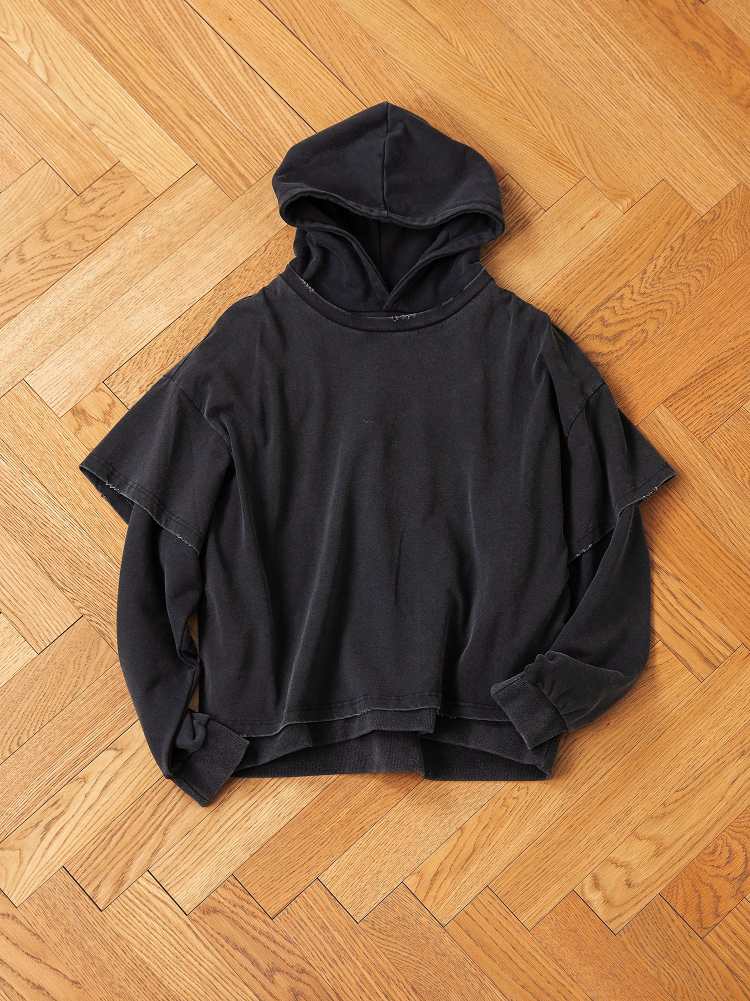 The Found Double Layer Hoodie, made from French terry cotton, is displayed on a wooden herringbone floor, highlighting its distinct design featuring layered short and long sleeves for a vintage appearance.