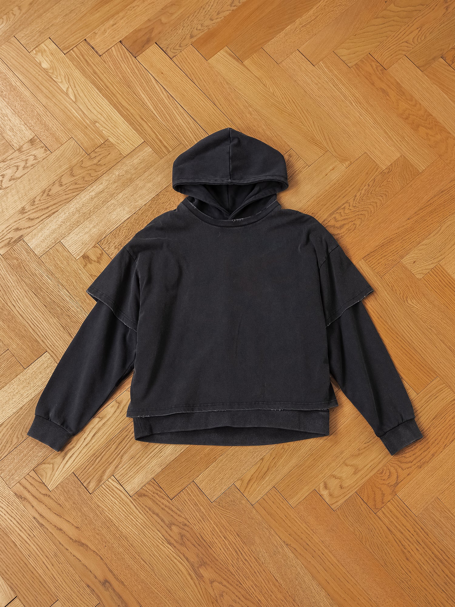 The Double Layer Hoodie by Found, featuring black long sleeves and crafted from French terry cotton, is laid flat on a wooden herringbone floor, exuding a vintage look.