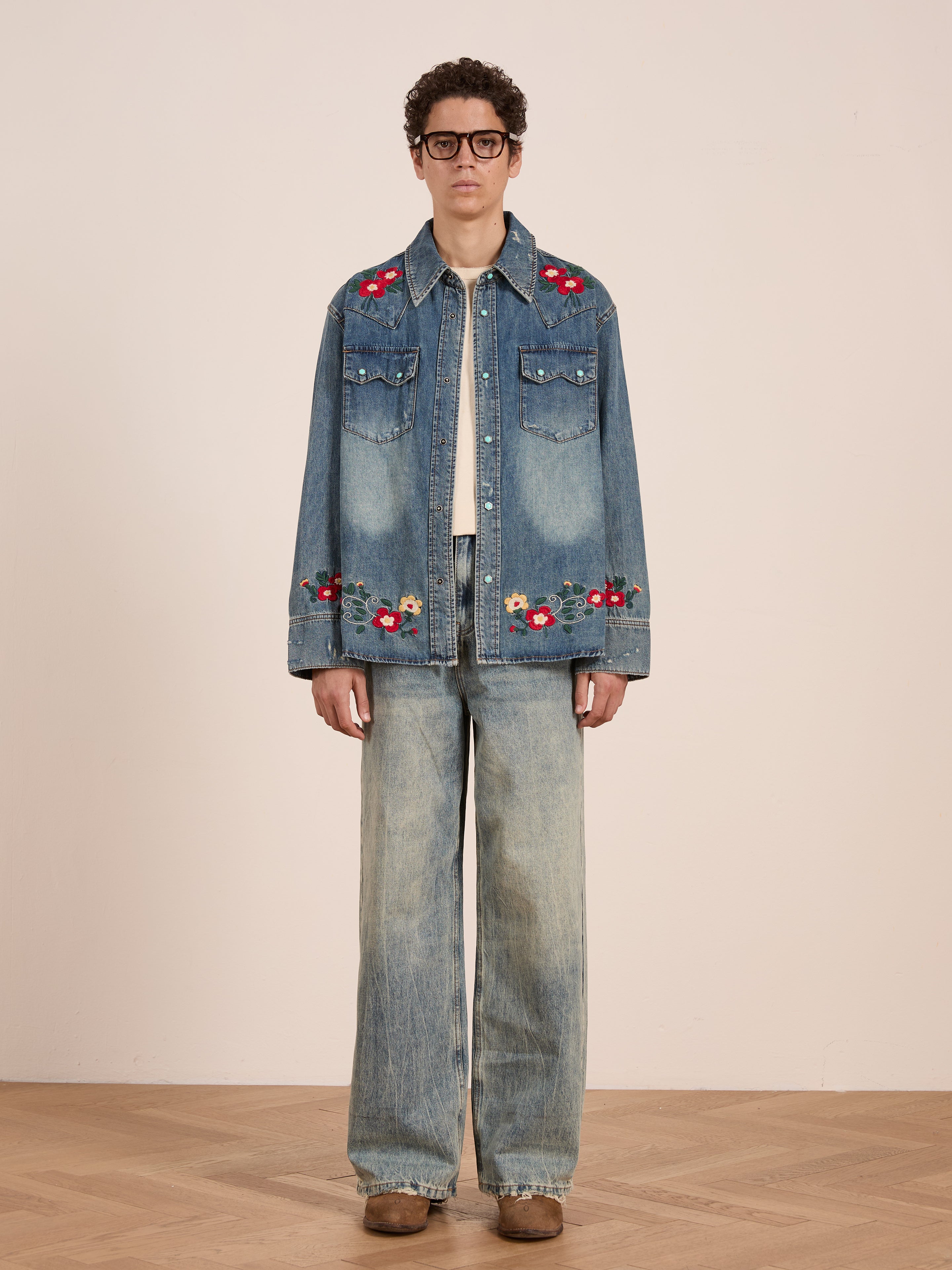 A person wearing glasses, the FOUND Floral Embroidered Western Denim Shirt, wide-leg jeans, and brown shoes stands against a plain light background.
