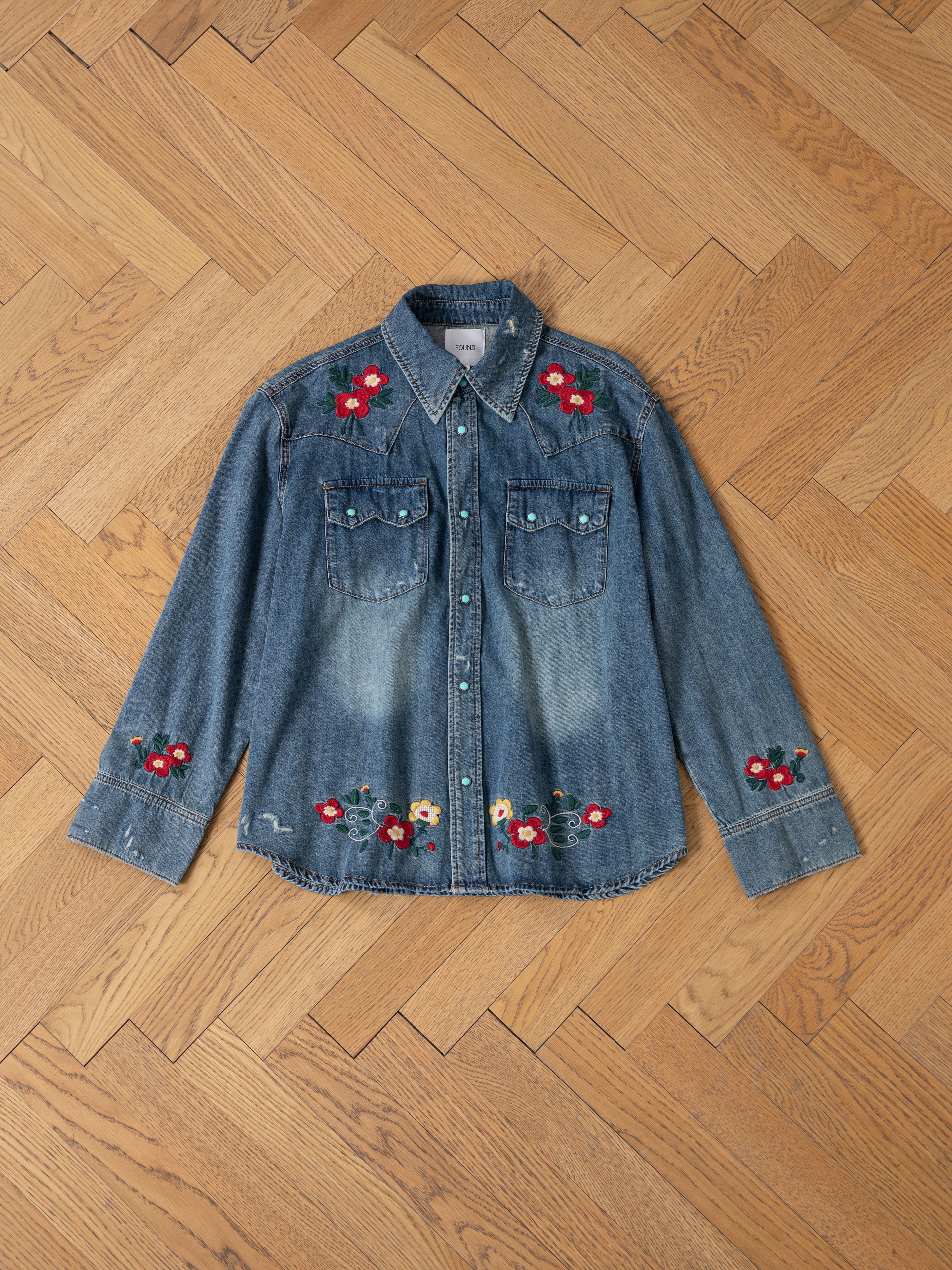 The FOUND Floral Embroidered Western Denim Shirt features red and yellow floral embroidery on the shoulders, front pockets, cuffs, and hem, shown on a light wooden herringbone floor.