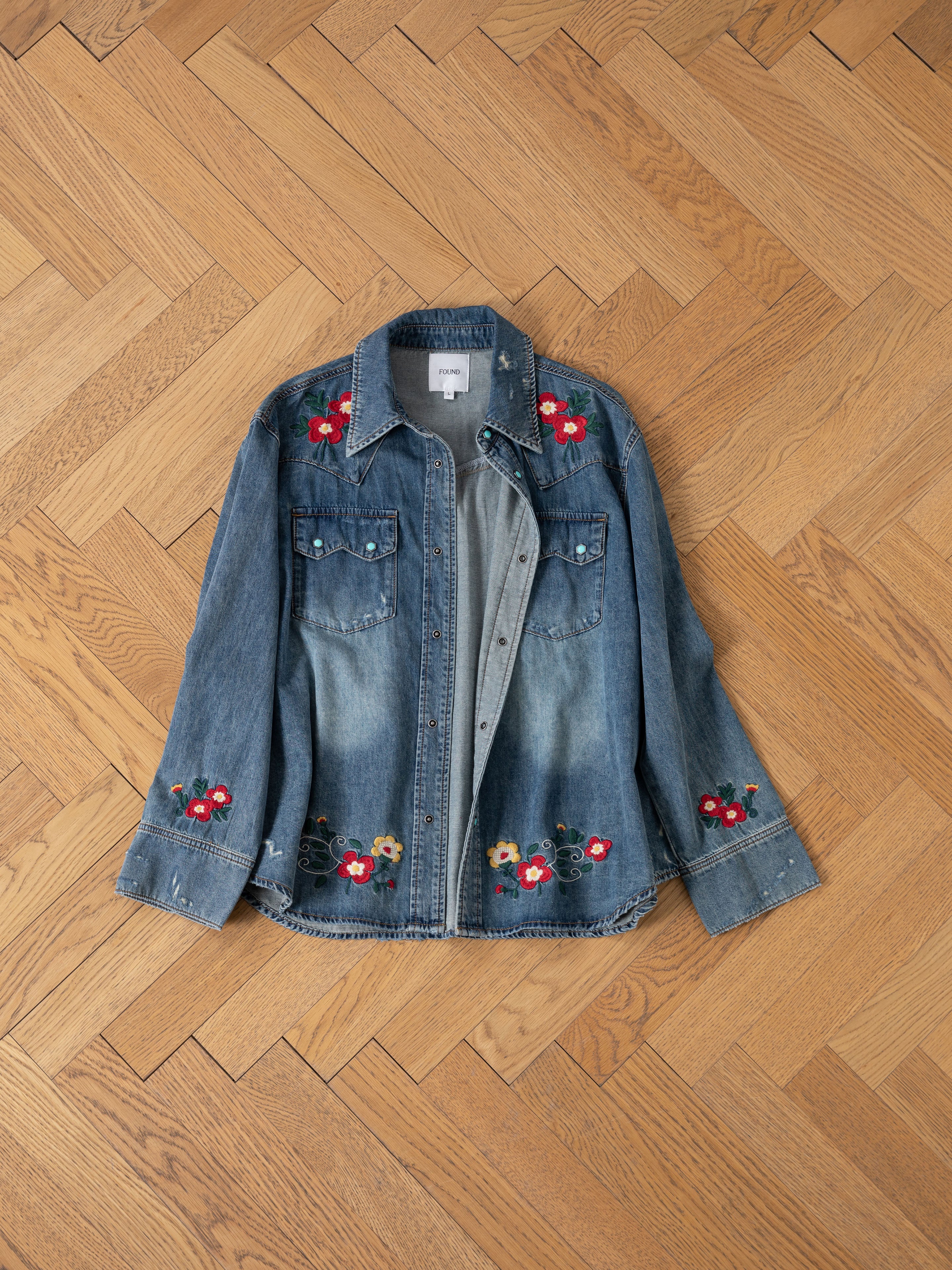 The FOUND Floral Embroidered Western Denim Shirt features chest pockets and delicate floral embroidery on the shoulders, cuffs, and hem, displayed flat on a wooden parquet floor.
