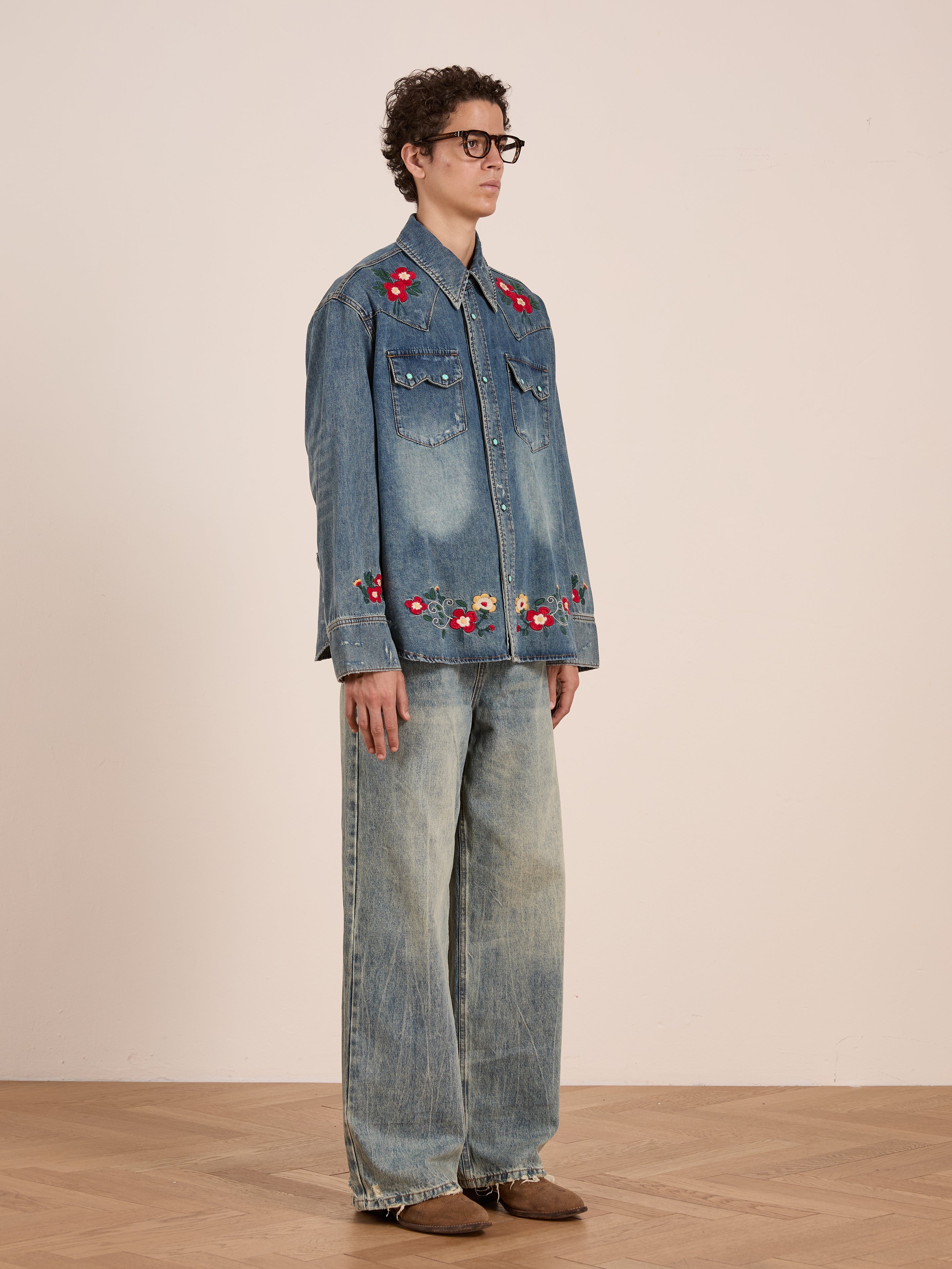 Someone wearing glasses and the FOUND Floral Embroidered Western Denim Shirt stands indoors against a plain wall, paired with wide-leg faded jeans and brown shoes.