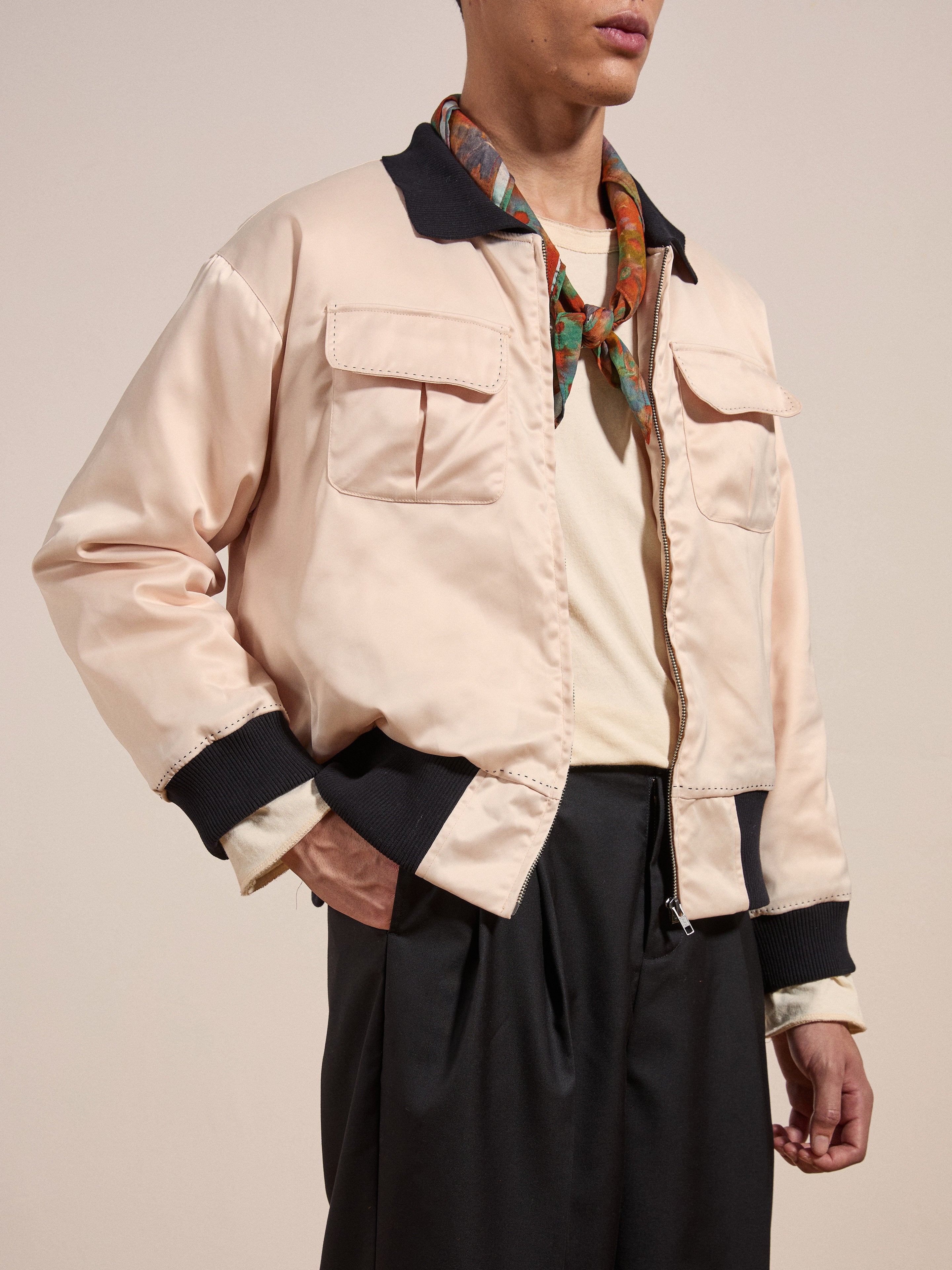 A person wearing a light pink Daman Satin Jacket by FOUND, accessorized with a green and orange scarf, paired with a white shirt and dark trousers, standing against a plain background.