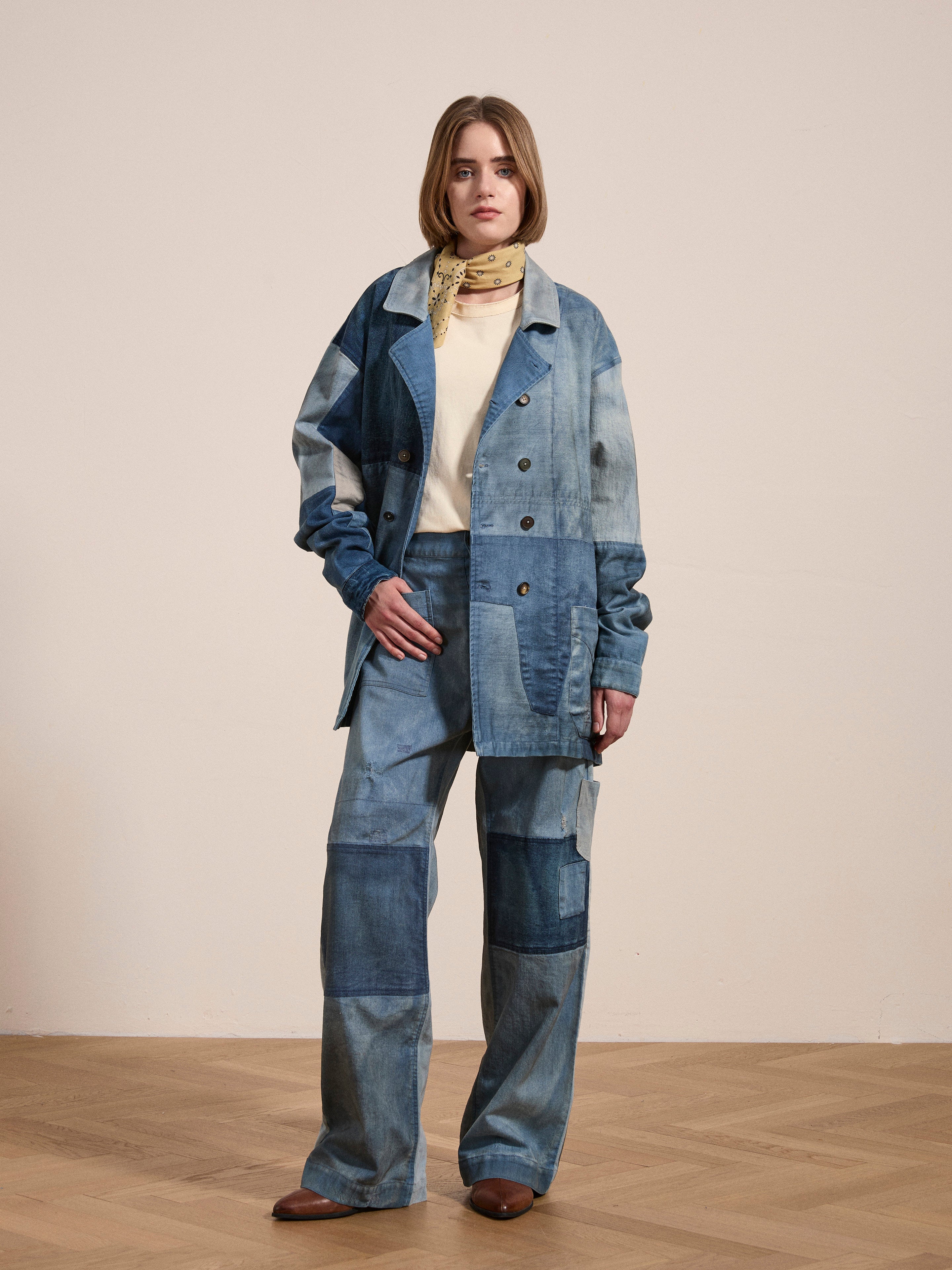 A person wearing a FOUND Multi Patch Painters Work Jacket with hand distressing, paired with patchwork denim pants, a light-colored turtleneck shirt, and a light scarf, is standing on a wooden floor against a plain background.