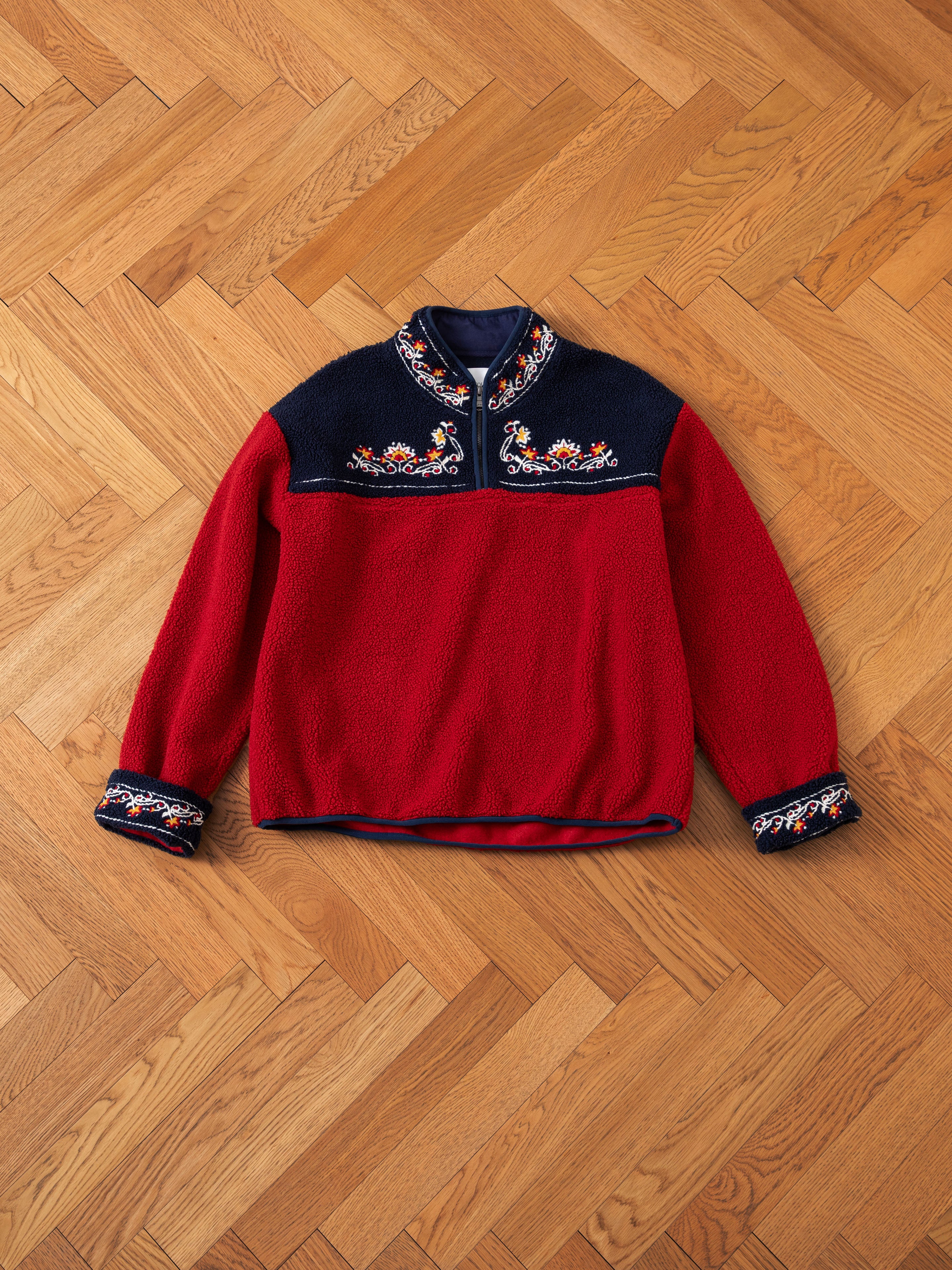 The FOUND Pahari Floral Embroidered Sherpa Pullover, a red and black long-sleeve sweater adorned with intricate Pahari style floral embroidery on the chest and cuffs, laid flat on a wooden floor with a herringbone pattern.