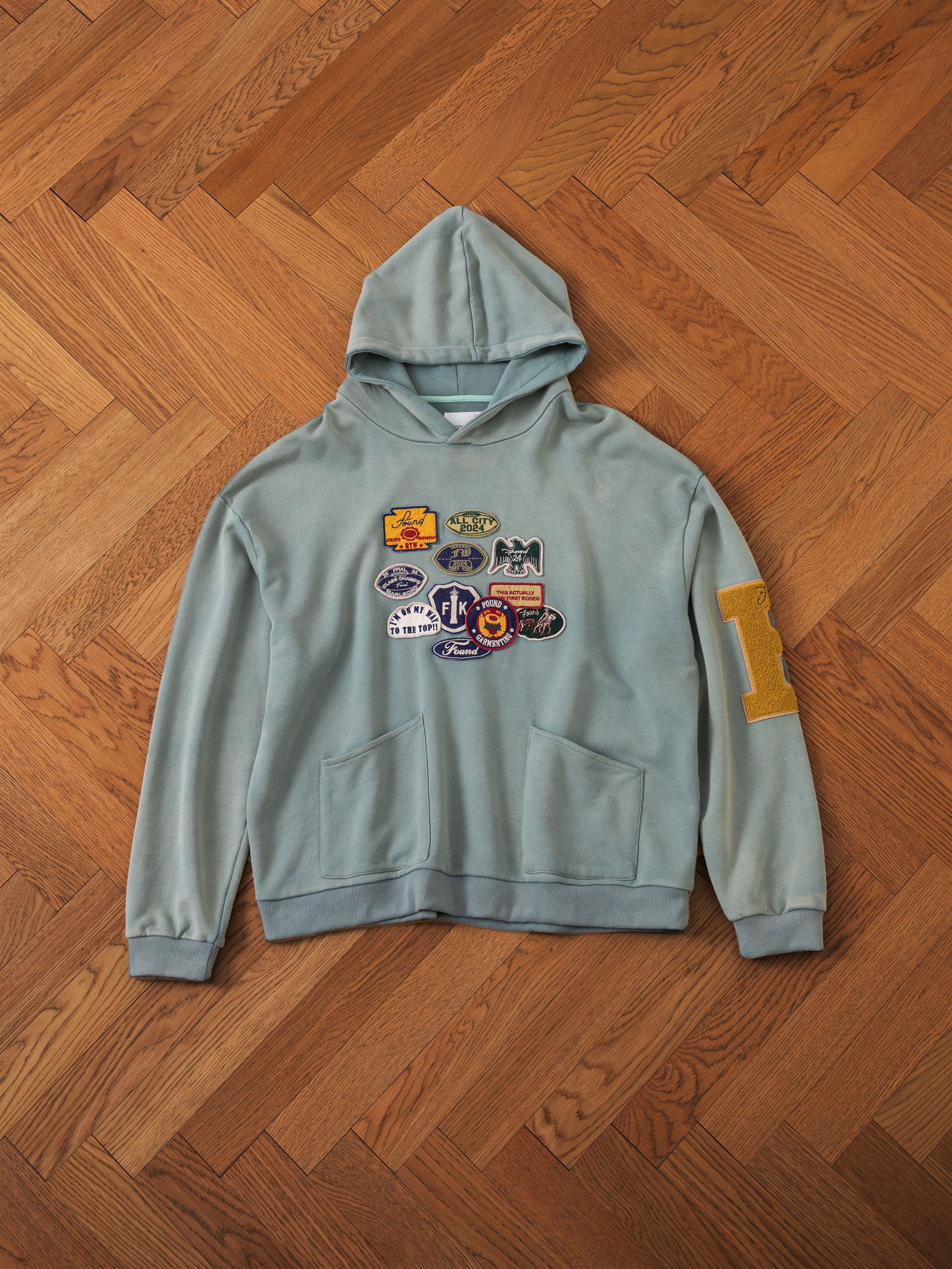 A light green Multi Patch Hoodie from FOUND, crafted from French Terry cotton and adorned with multiple colorful patches on the front and a large yellow "R" patch on the left sleeve, is laid out on a wooden parquet floor.