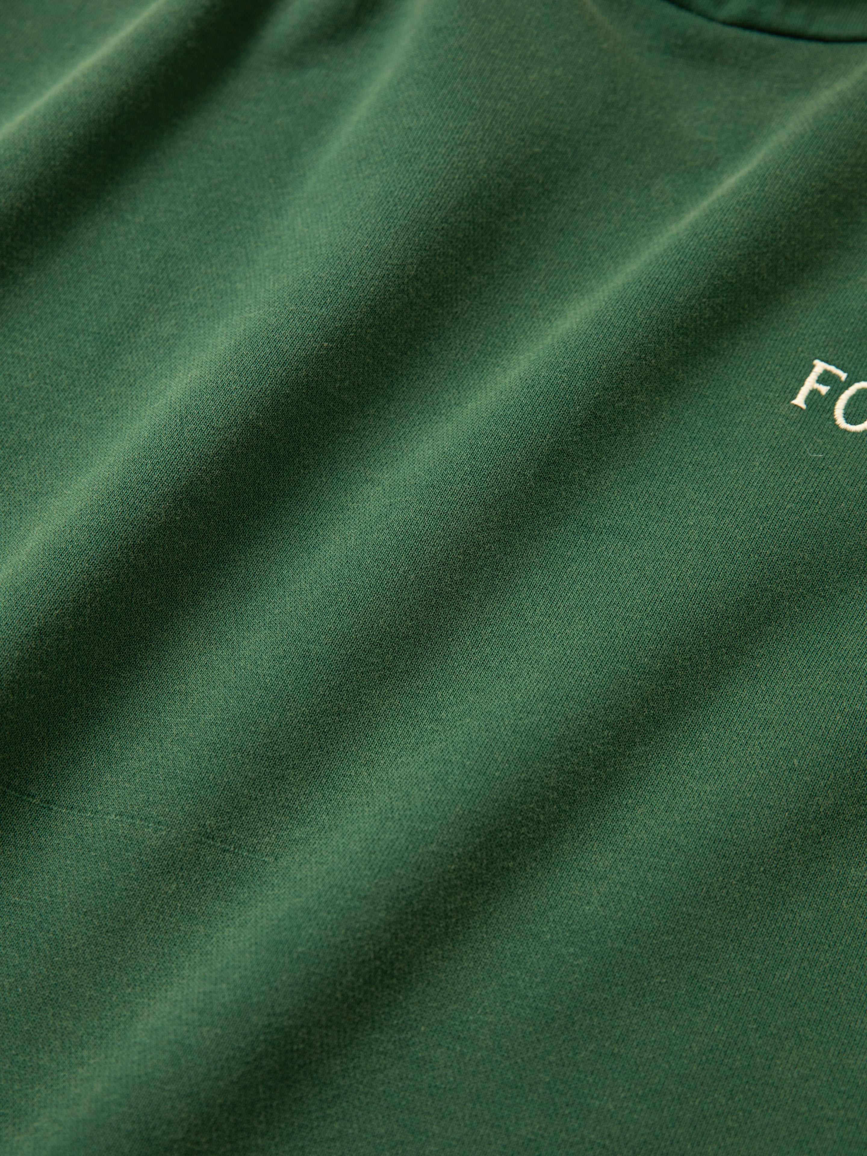 Close-up of the upper section of a green Stitched Border Distressed Logo Hoodie by FOUND, with a small portion of white text visible, reading 'FO'. The fabric appears smooth and evenly colored, reminiscent of the classic vintage feel found in oversized hoodies.