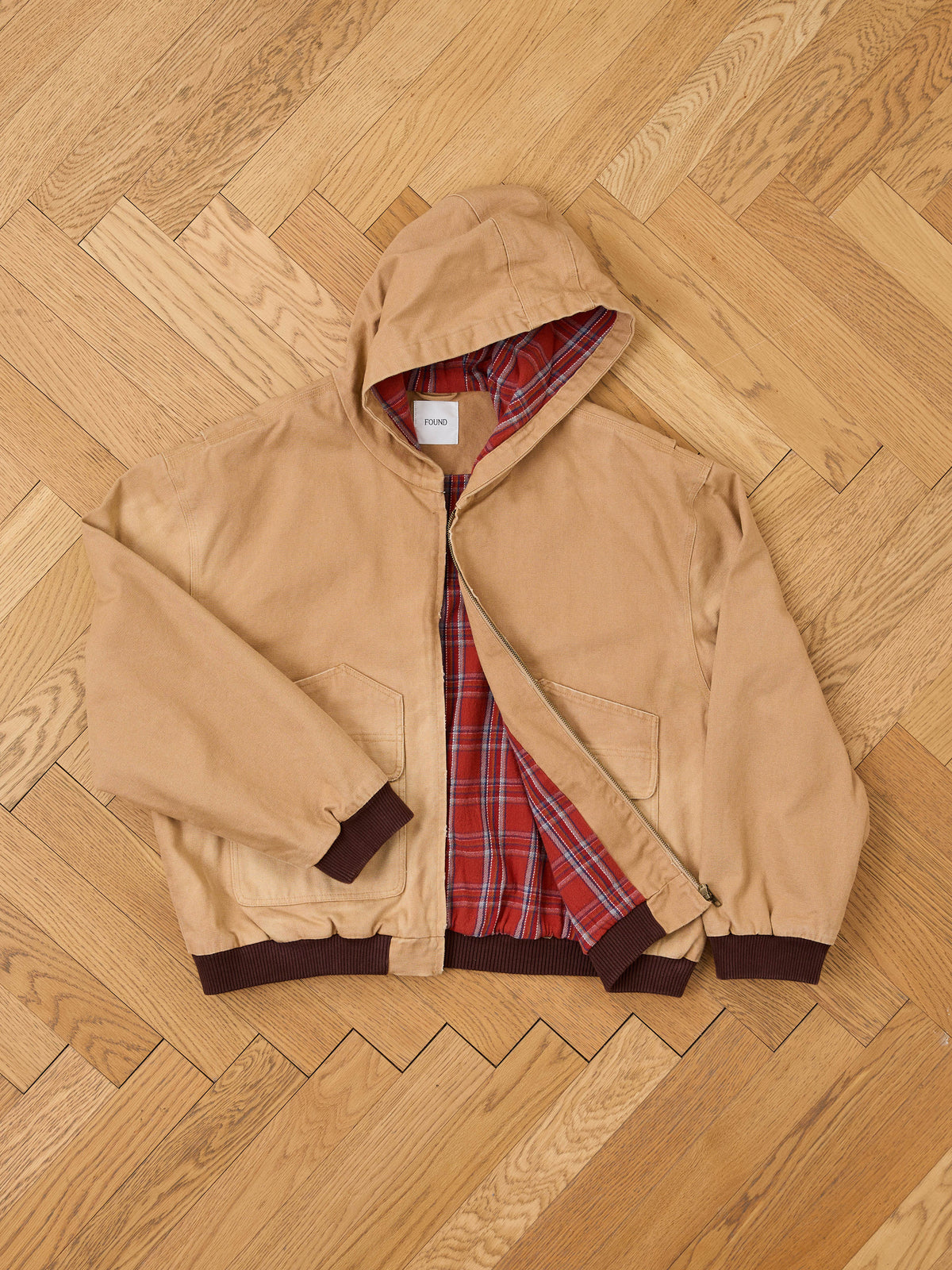 The FOUND Faded Canvas Work Hoodie features a tan canvas body, brown cuffs and hem, and a red plaid lining for unisex relaxed comfort. Displayed unzipped on a wooden floor.