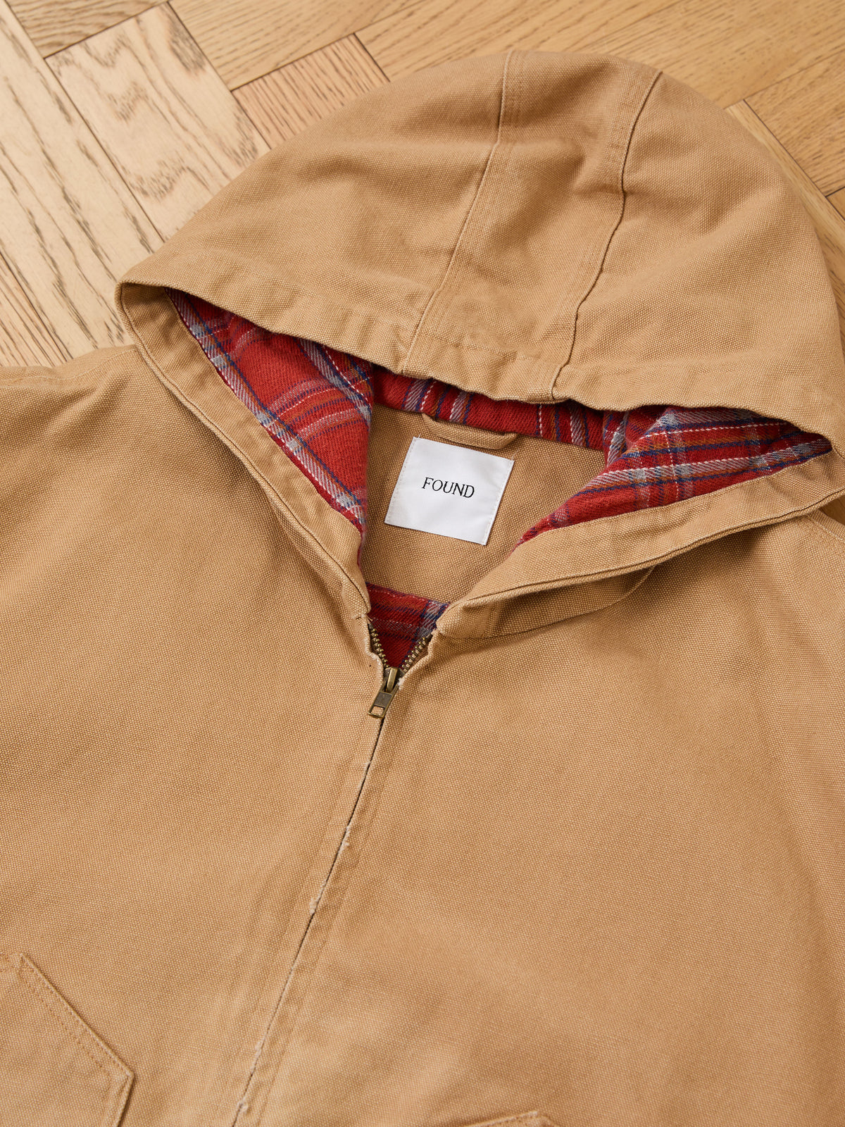 The Faded Canvas Work Hoodie by FOUND, tan with red plaid lining and unisex relaxed fit, is shown laid flat on a wooden floor.