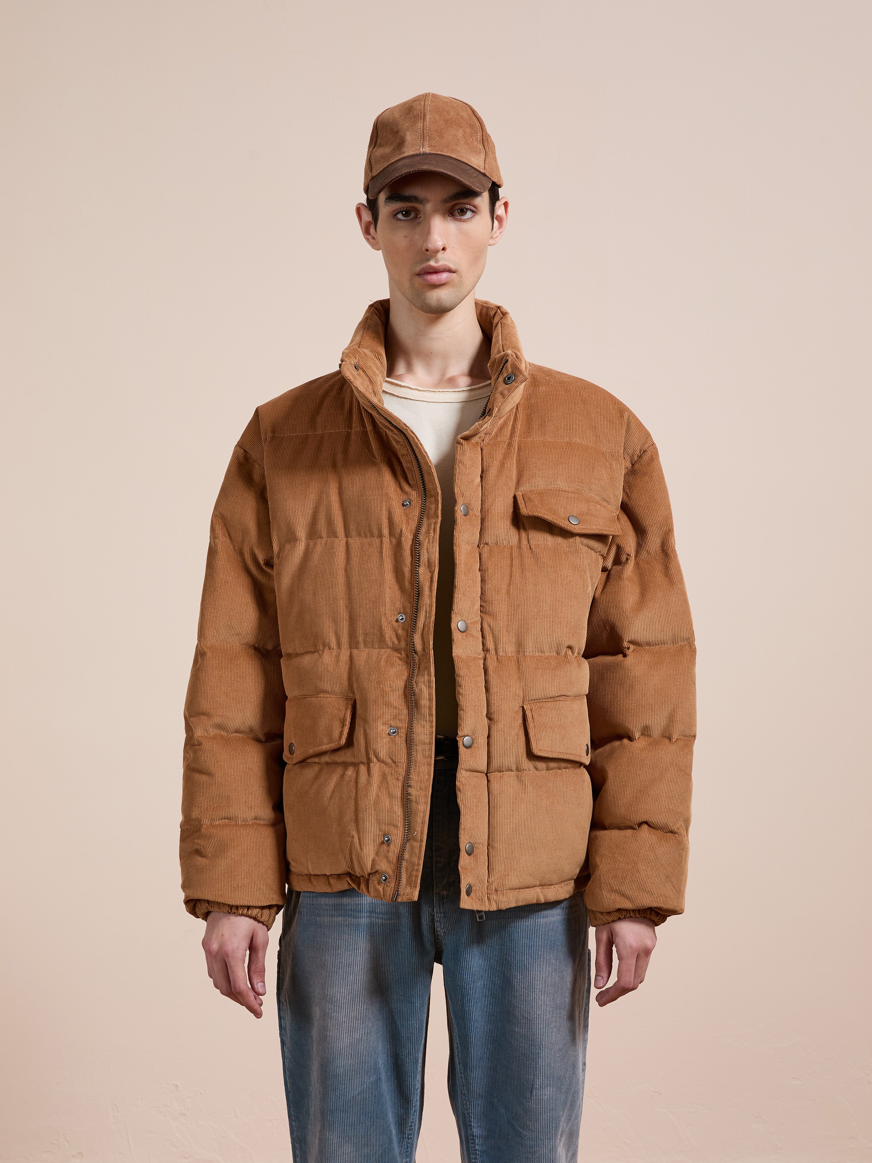 A person stands against a beige backdrop wearing the FOUND Corduroy Puffer Jacket in tan with a relaxed fit, paired with a matching tan baseball cap, light shirt, and blue jeans.