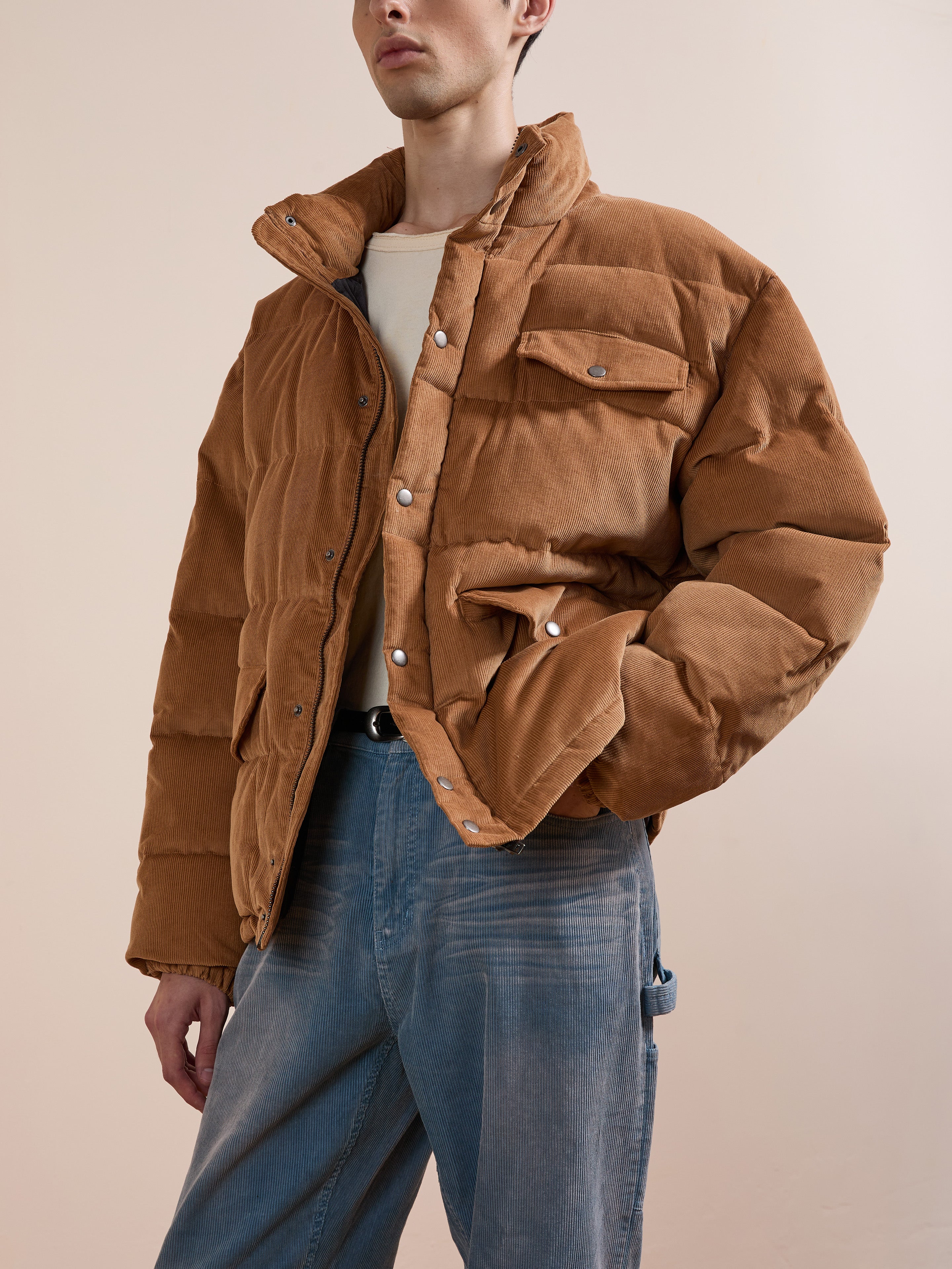 A person models the FOUND Corduroy Puffer Jacket in tan, styled with a light shirt and blue jeans, standing against a plain beige background.