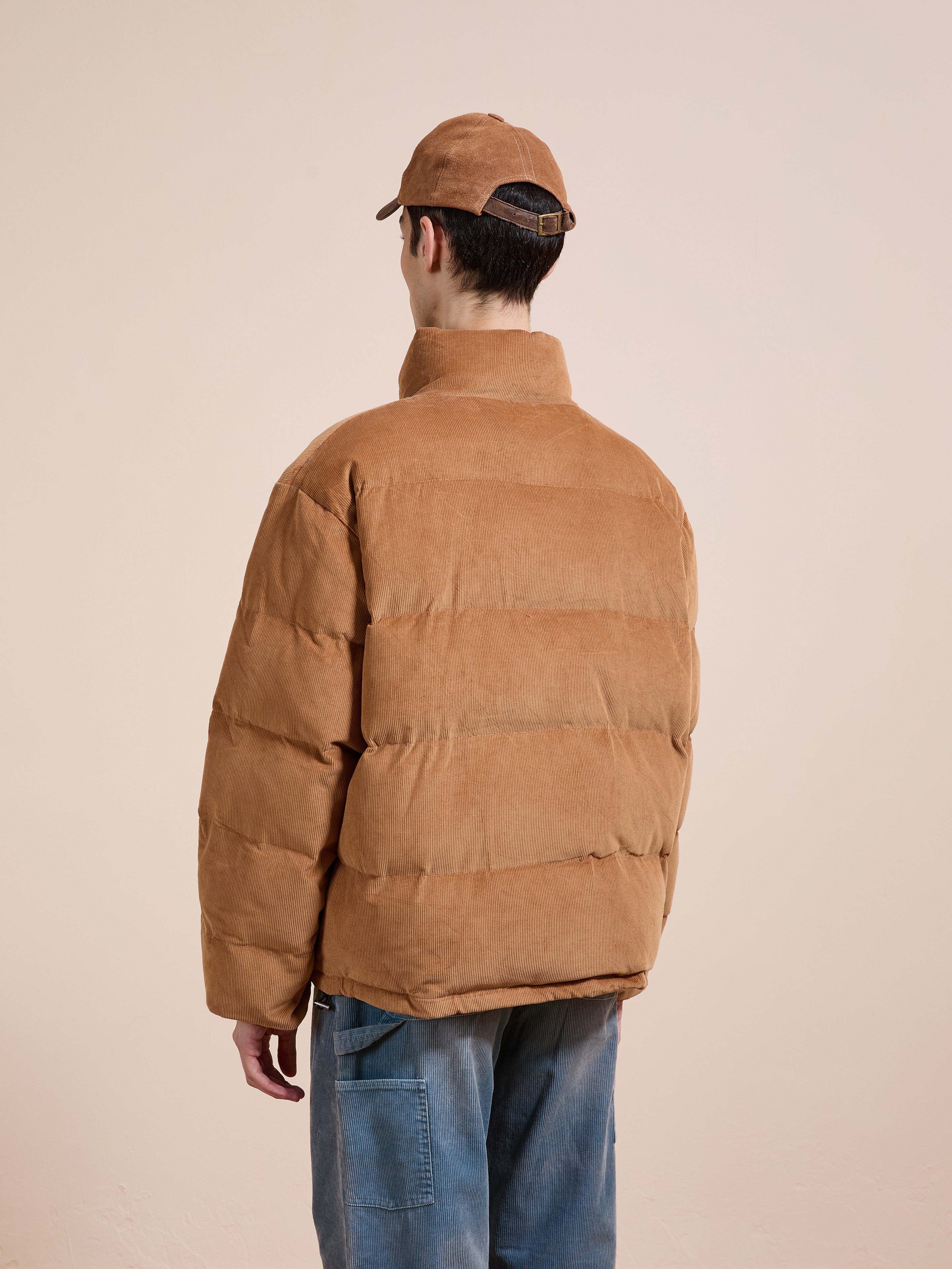 A person wearing the FOUND Corduroy Puffer Jacket in tan, a tan cap, and blue jeans stands facing away from the camera against a plain light background.