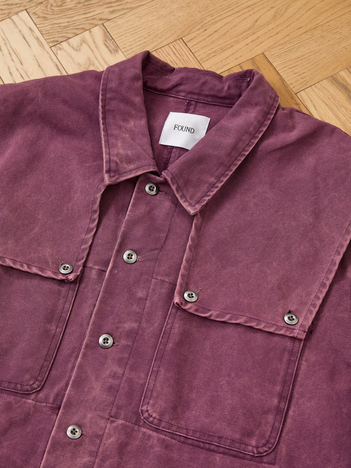A mauve Faded Paneled Cargo Jacket by Found with two chest pockets lies flat on a wooden floor. The visible label inside the collar reads “Found.” This unisex relaxed fit piece channels classic utility outerwear, making it both versatile and effortlessly stylish.