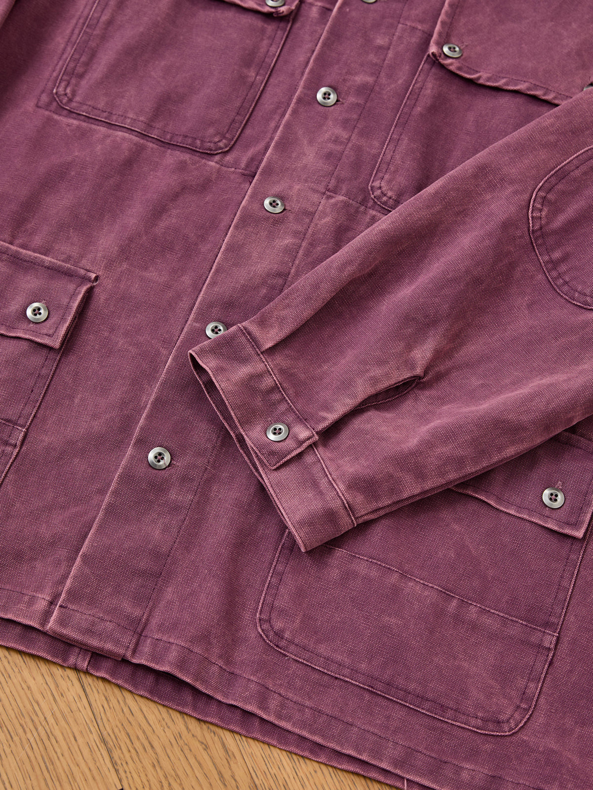 Close-up of the Found Faded Paneled Cargo Jacket in mauve, featuring large front pockets, metal snap buttons, and visible stitching, laid flat on a wooden surface. This utility outerwear offers a unisex relaxed fit and highlights the distinctive look of its garment-dyed finish.