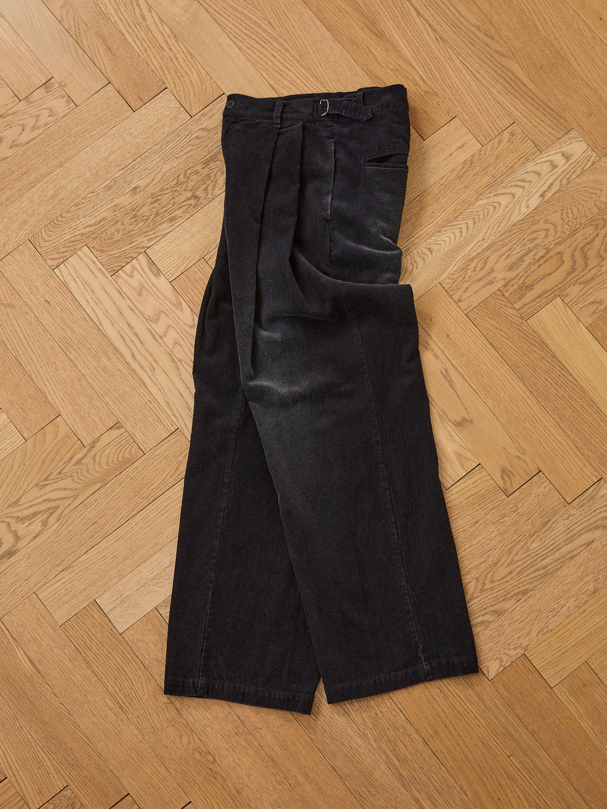A pair of FOUND Faded Pleated Corduroy Pants is displayed flat on a light wooden herringbone floor.