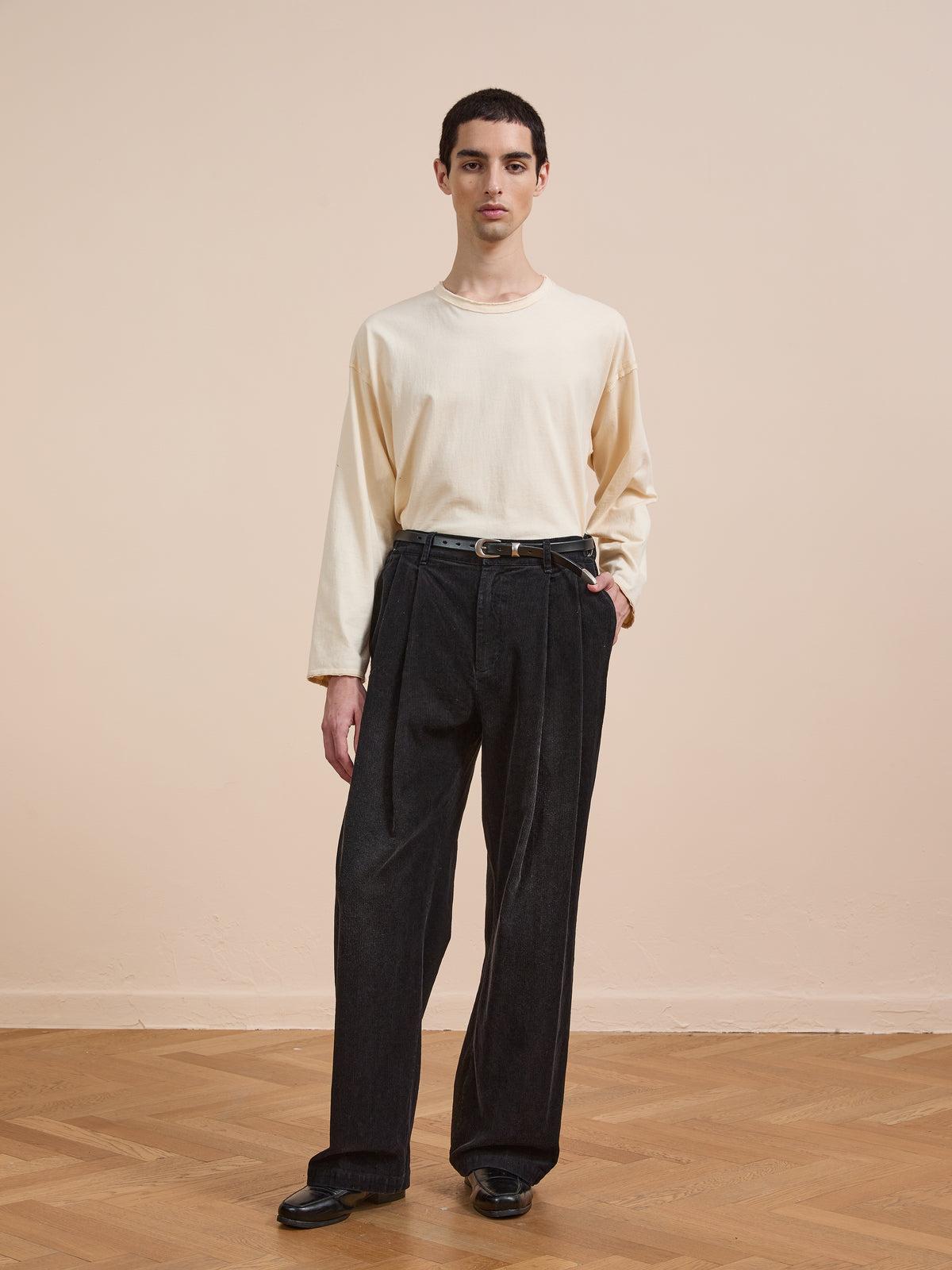 A person stands indoors on a wooden floor, wearing FOUND’s Faded Pleated Corduroy Pants paired with a long-sleeved cream shirt, black shoes, and a belt, set against a plain beige background.