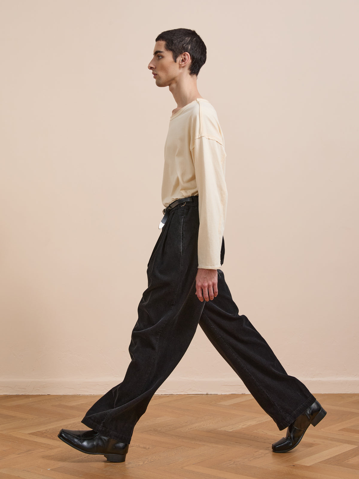 A person with short dark hair walks indoors on a wooden floor, wearing a beige long-sleeve shirt, black shoes, and FOUND Faded Pleated Corduroy Pants.