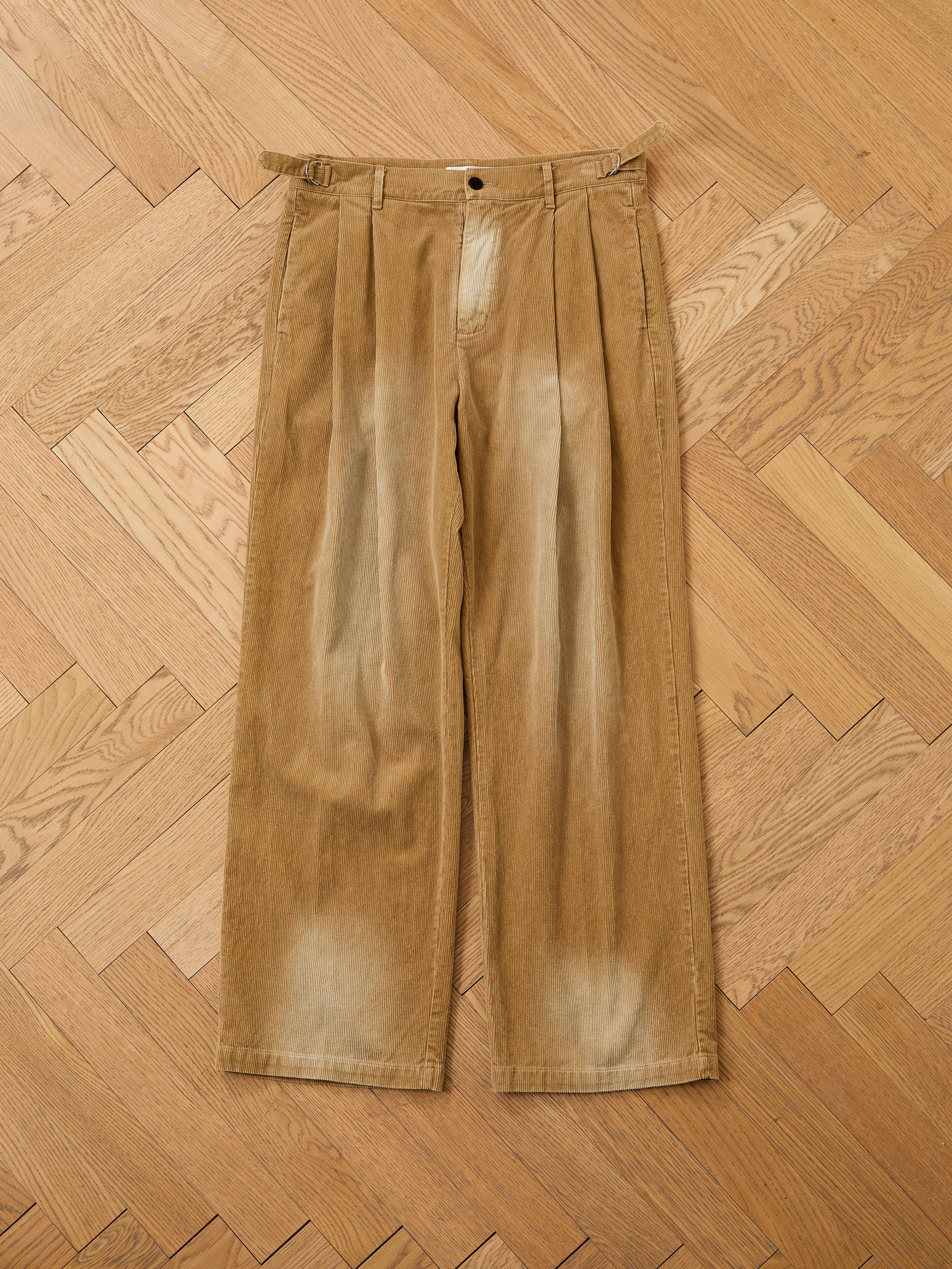 FOUND Faded Pleated Corduroy Pants in tan, featuring a relaxed wide-leg fit with pleats and faded details on the thighs and hems, shown flat on a wooden parquet floor.