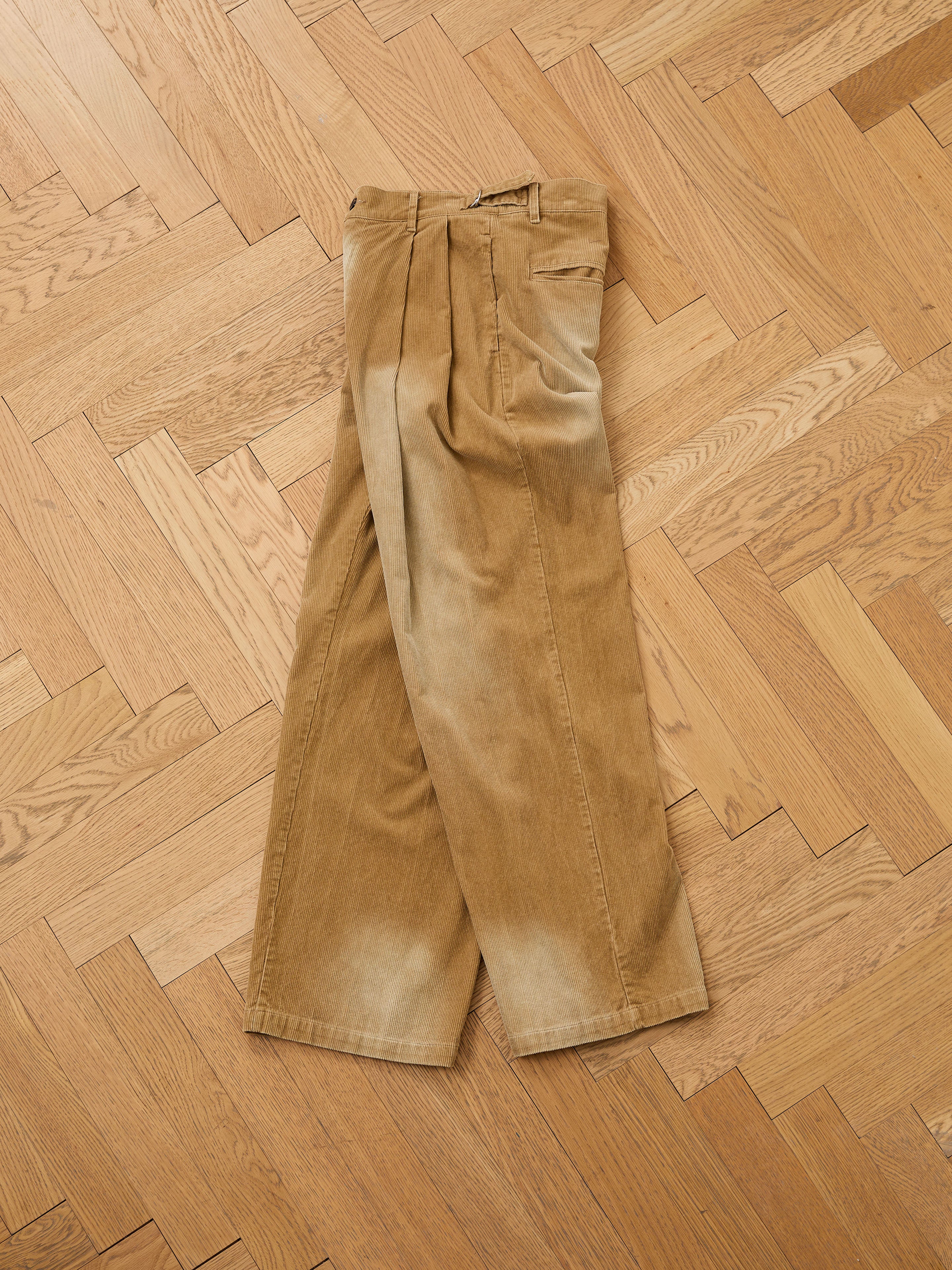 The FOUND Faded Pleated Corduroy Pants, featuring a relaxed fit and double pleats, are folded neatly on a wooden parquet floor.