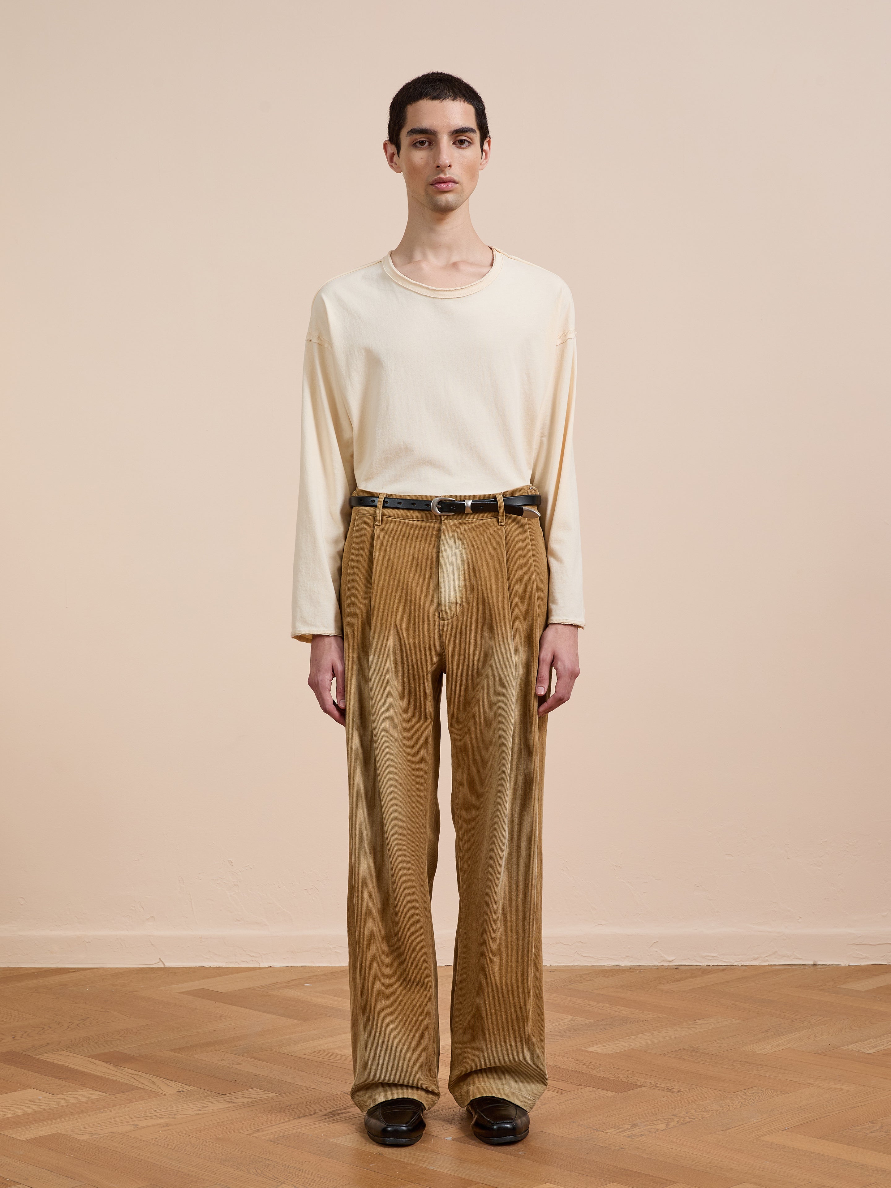 A person stands indoors against a beige wall, wearing a cream long-sleeve shirt, FOUND’s Faded Pleated Corduroy Pants, a black belt, and black shoes.