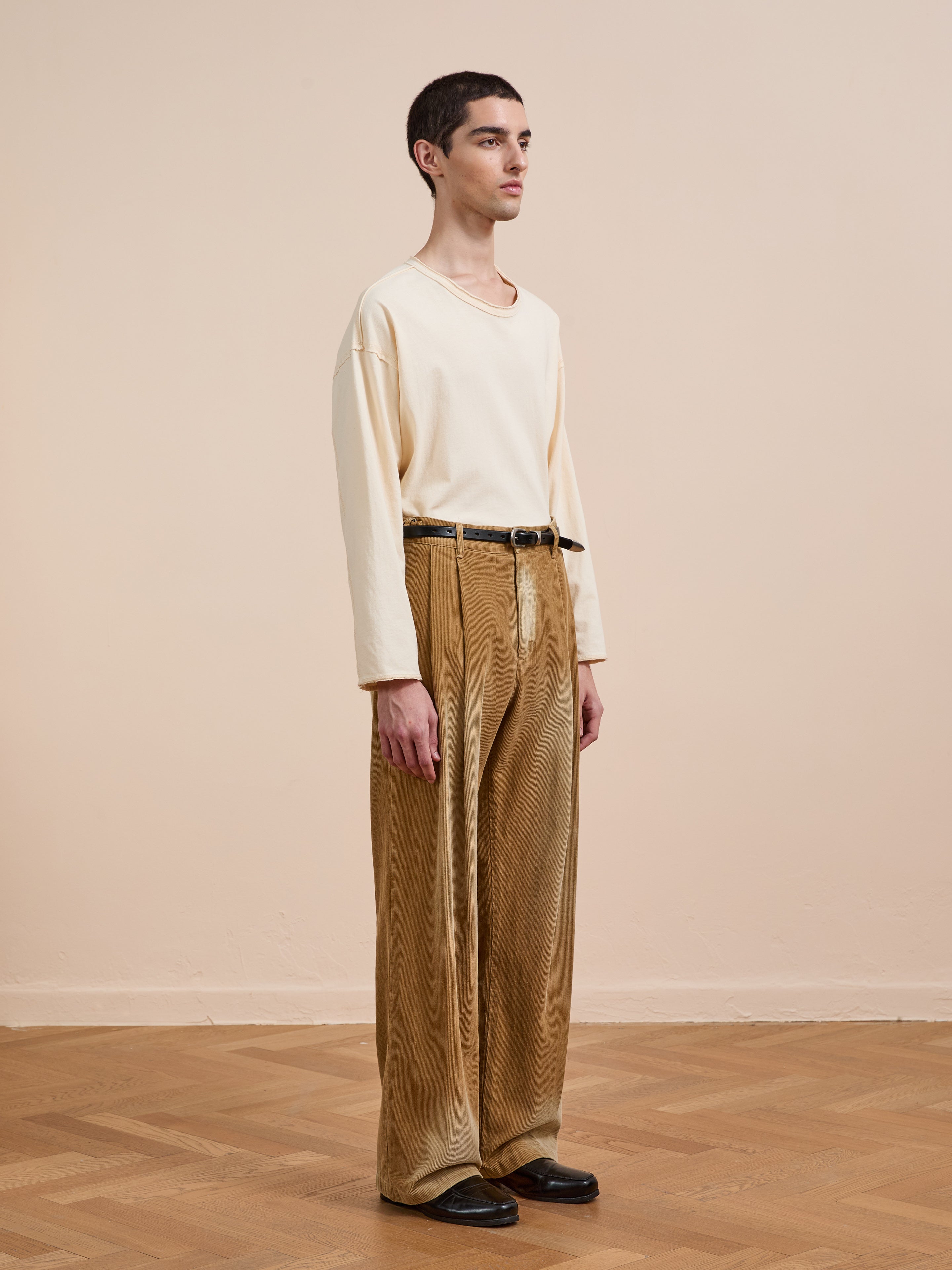 Indoors on a parquet floor, a person wears a loose beige long-sleeve shirt, black shoes, and FOUND’s Faded Pleated Corduroy Pants in tan, standing slightly turned to the side.