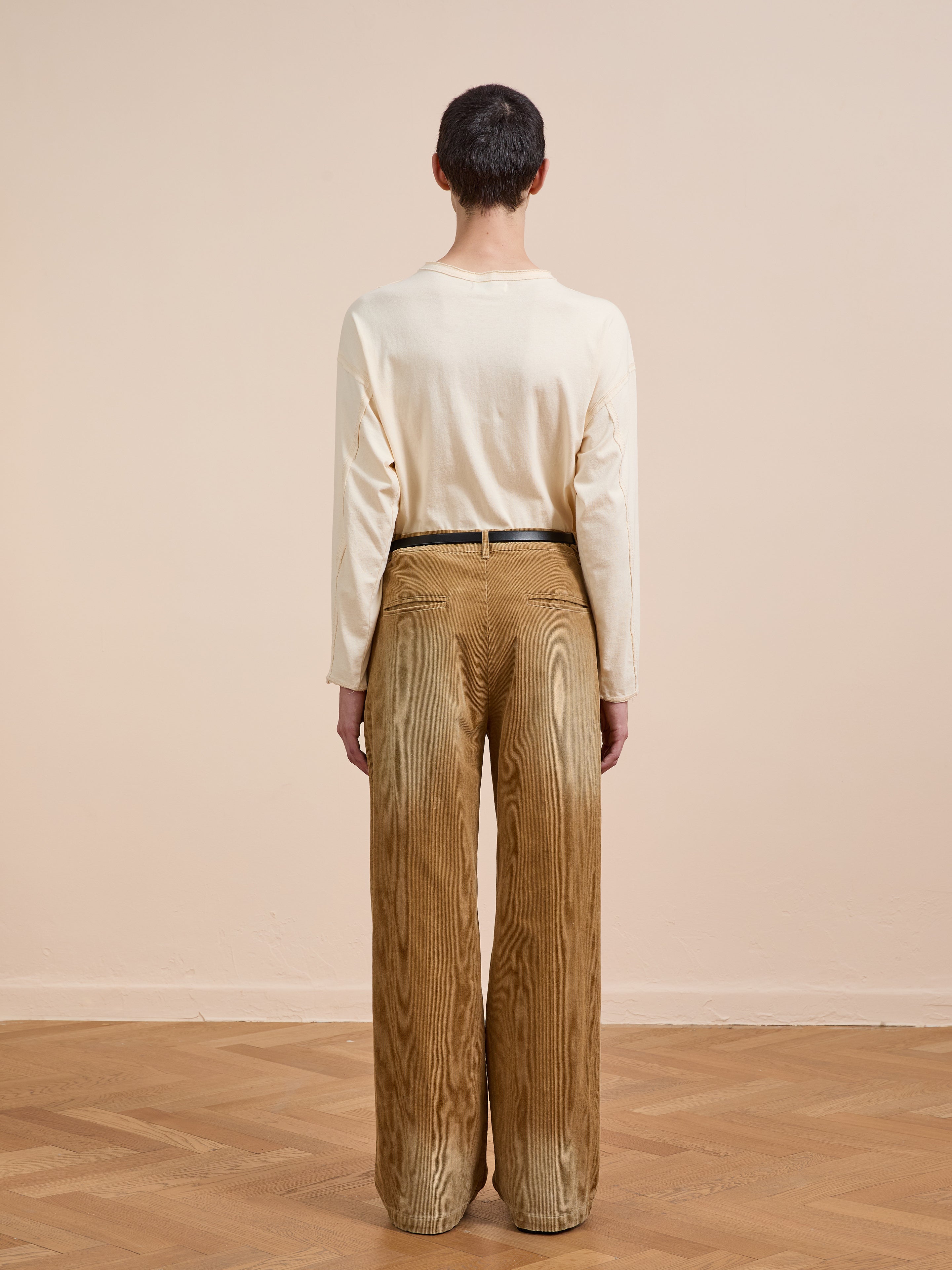 A person stands on a wooden floor facing away, wearing FOUND’s Faded Pleated Corduroy Pants with a long-sleeve cream shirt against a neutral background.