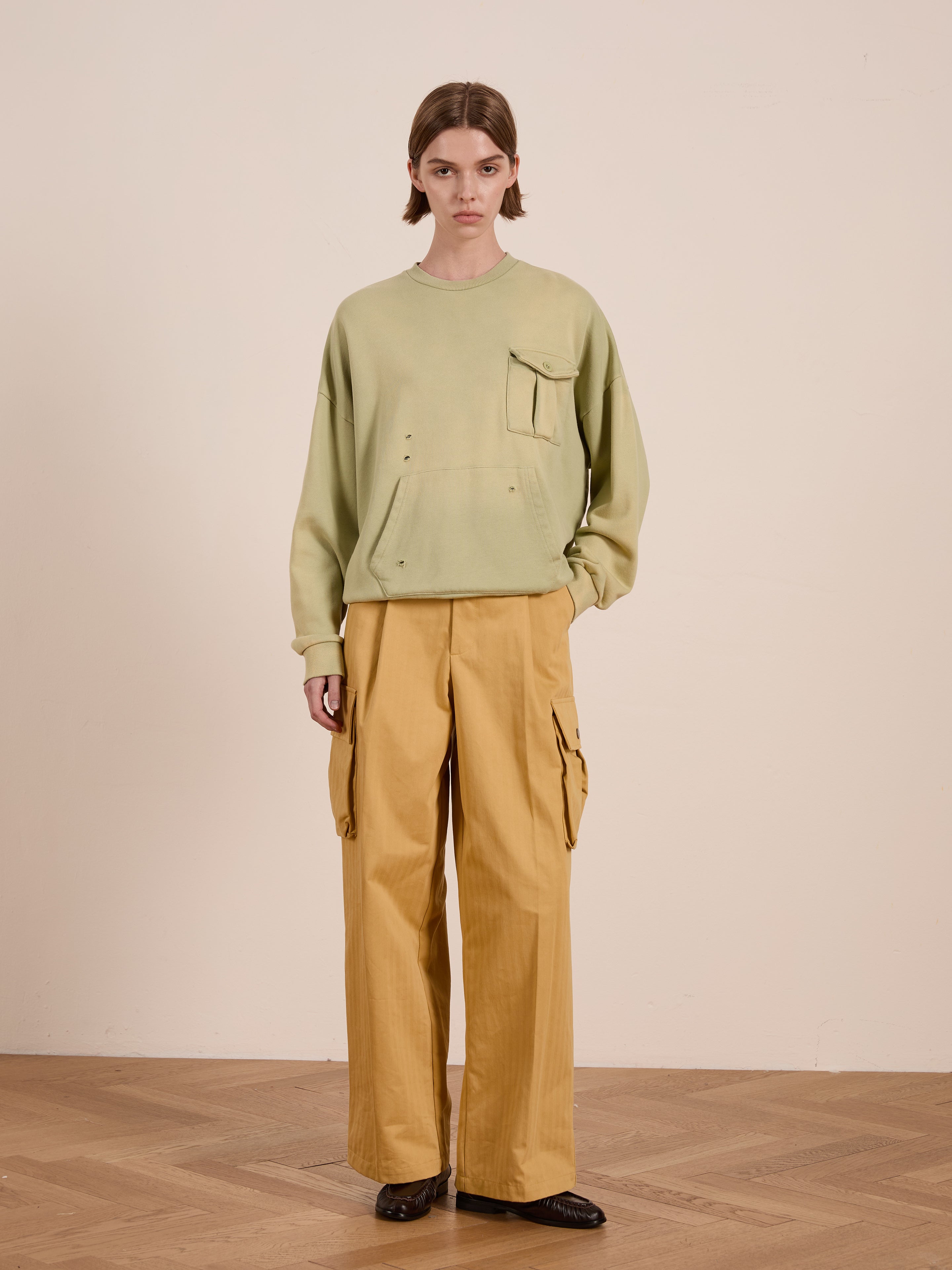 Wearing a light green Faded Pocket Crewneck by FOUND with its vintage-inspired fade and mustard yellow cargo pants, the individual captures the unisex relaxed fit look against a plain beige wall on a wooden floor.