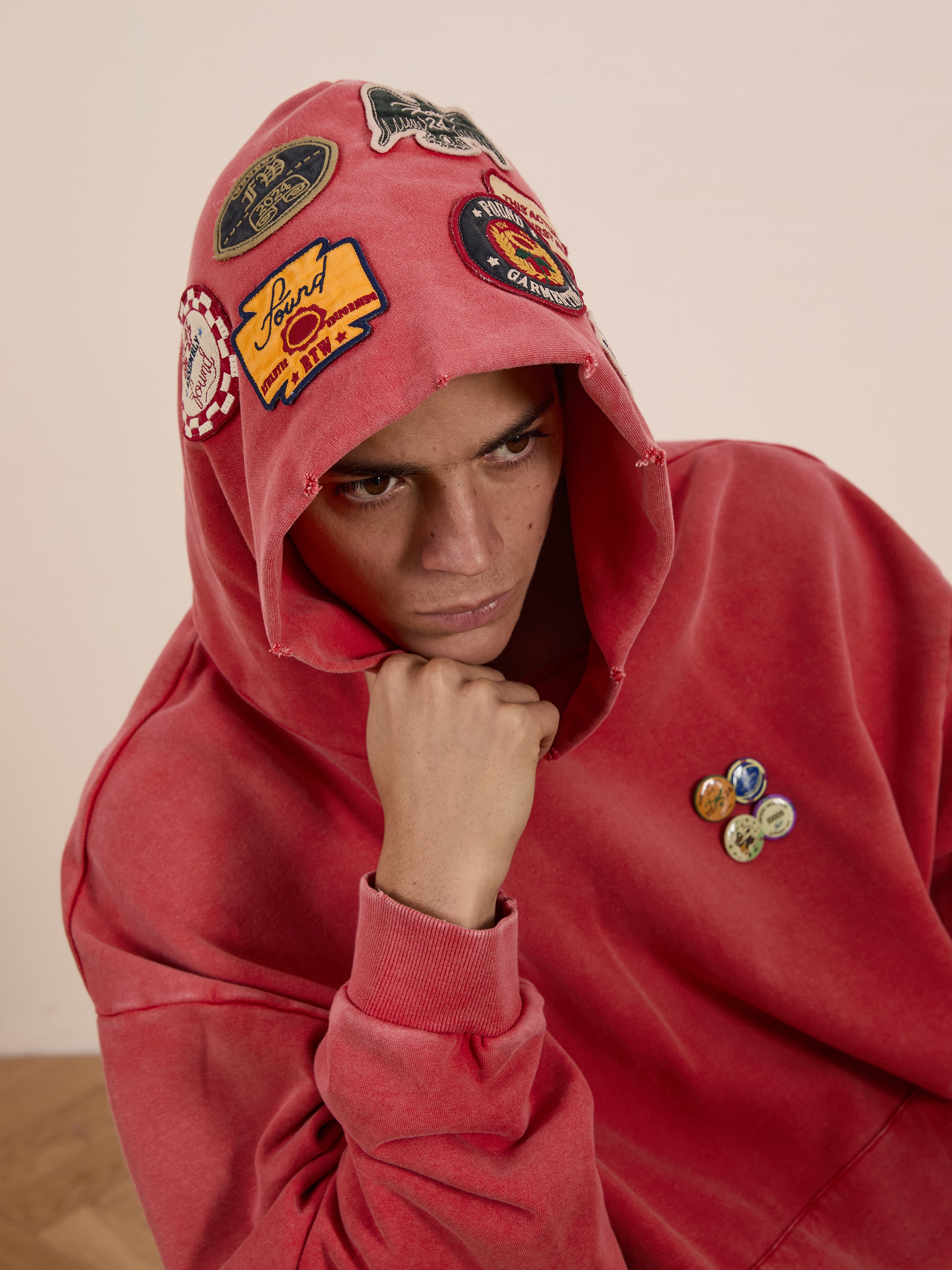 A person in a FOUND Multi Patch Hoodie, oversized and adorned with various patches and pins, rests their chin on their hand, appearing thoughtful.