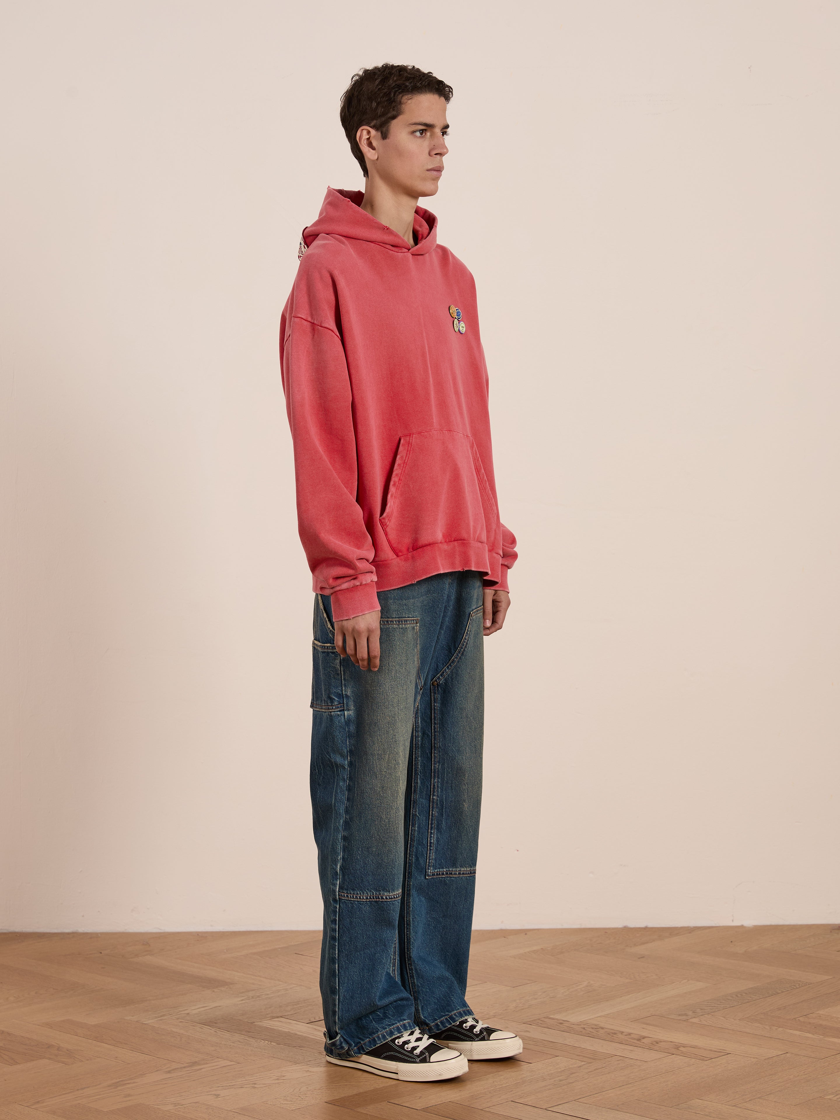 Someone wearing a FOUND Multi Patch Hoodie and wide-leg, sun-faded wash jeans stands on a wooden floor, facing left.