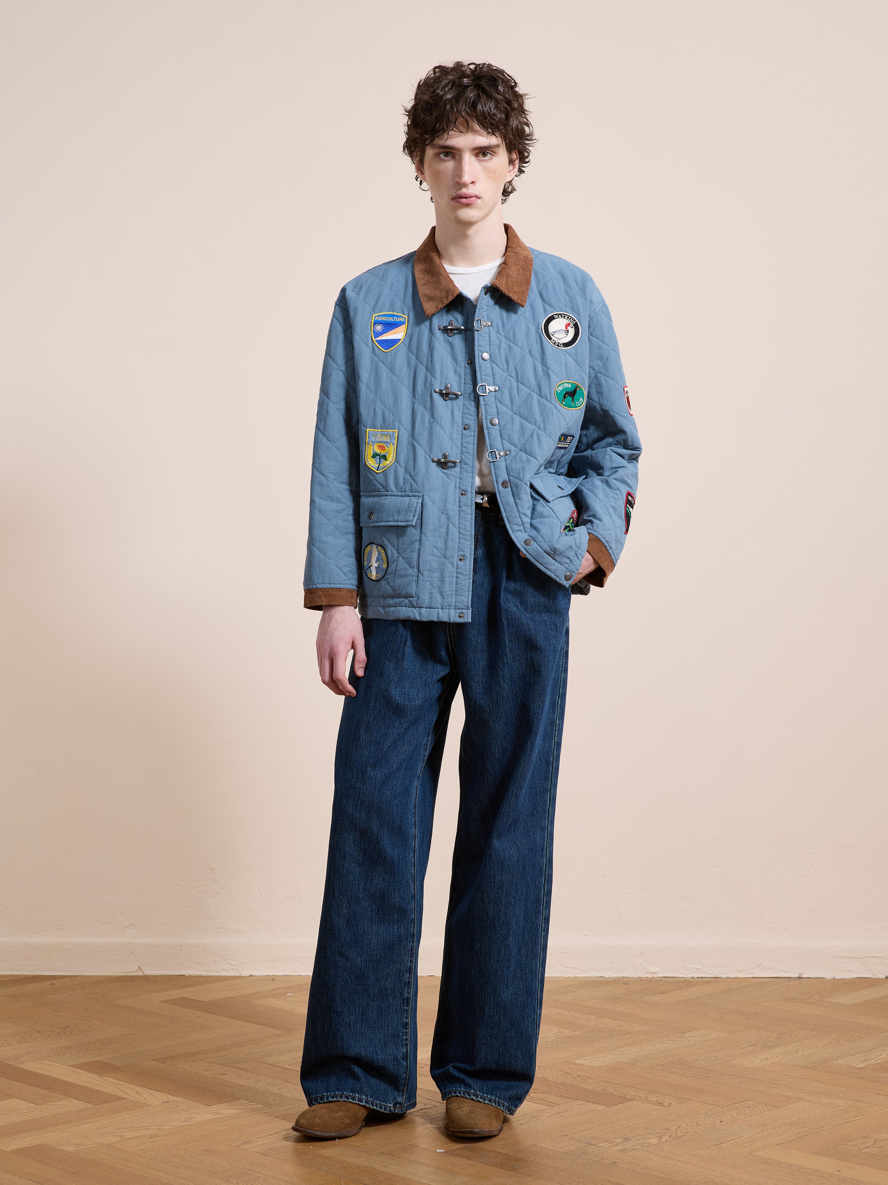 A person stands indoors against a plain background wearing the FOUND Farmstead Quilt Patch Jacket, wide-leg blue jeans, tan shoes, and a white shirt.