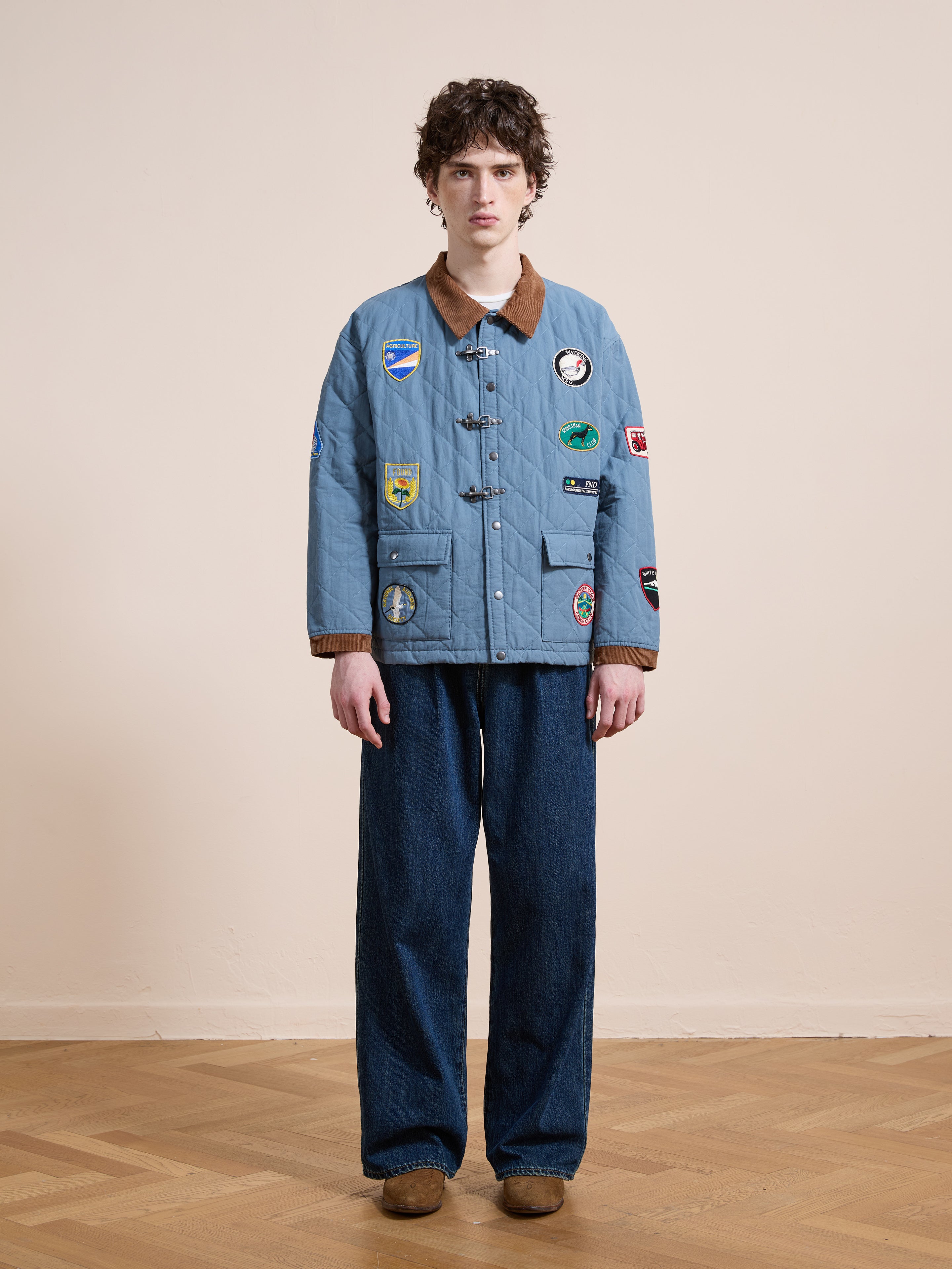 A person stands indoors against a beige wall and wooden floor, wearing the FOUND Farmstead Quilt Patch Jacket in blue with patches, styled with wide-leg jeans and brown shoes.