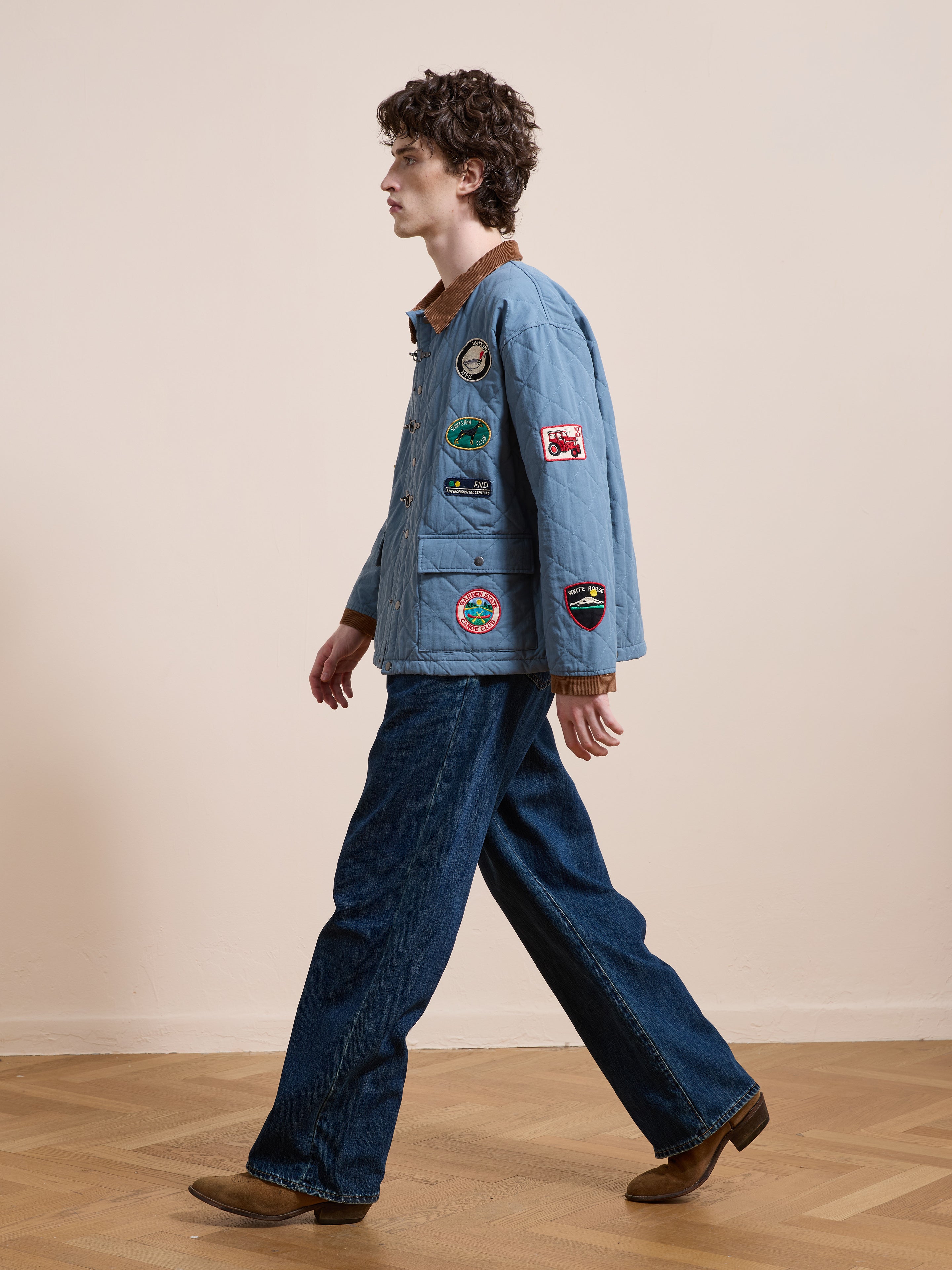 A person walks indoors on a wooden floor, wearing FOUND’s Farmstead Quilt Patch Jacket, wide-leg blue jeans, and brown shoes.