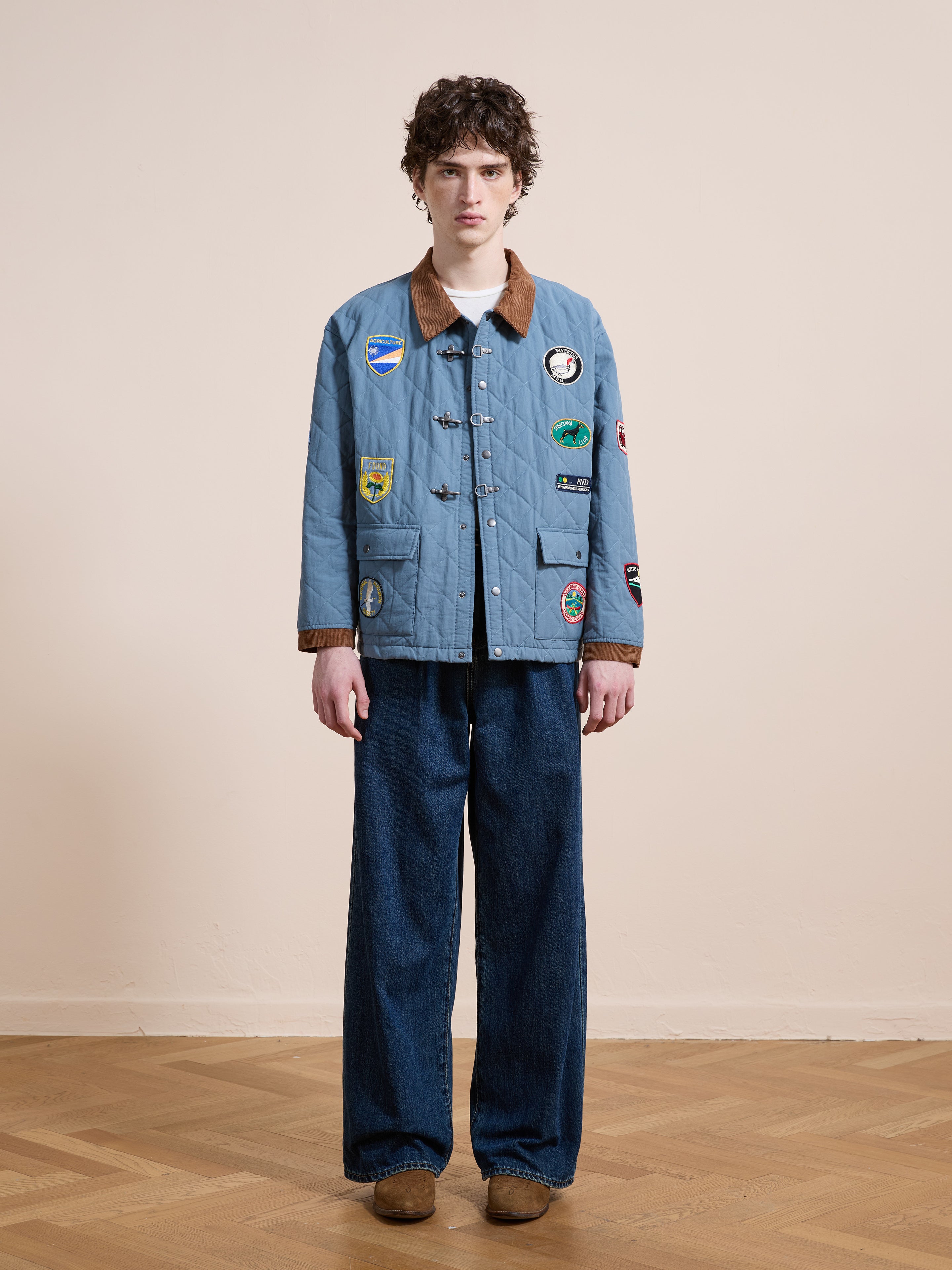 Indoors against a beige wall and wooden floor, a person wears the FOUND Farmstead Quilt Patch Jacket with wide-leg jeans and brown shoes.