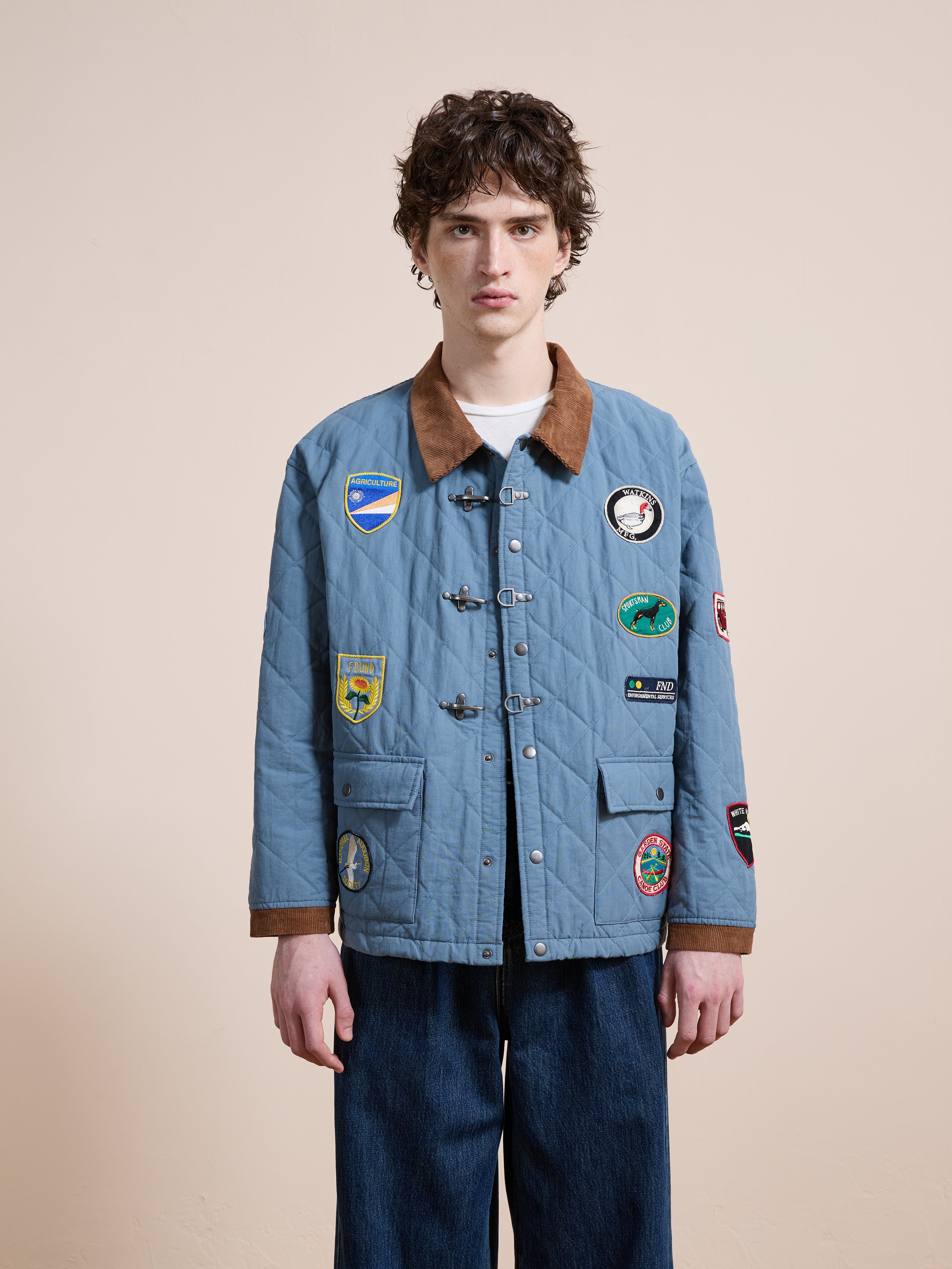 A person stands facing forward in the FOUND Farmstead Quilt Patch Jacket—light blue with a brown collar and colorful patches—paired with blue jeans, set against a plain beige background.