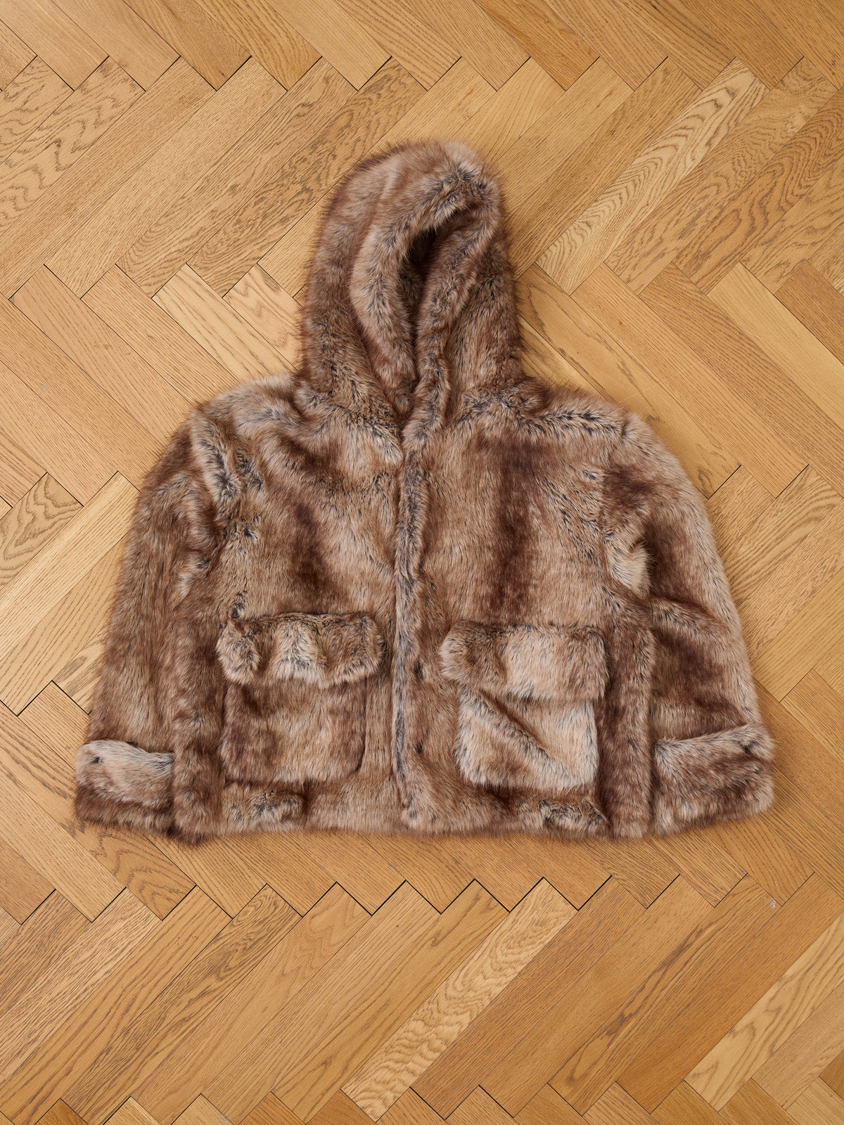 The FOUND Faux Fur Hoodie, a unisex brown jacket with two front pockets, is displayed flat on a herringbone floor for a vintage outerwear vibe.