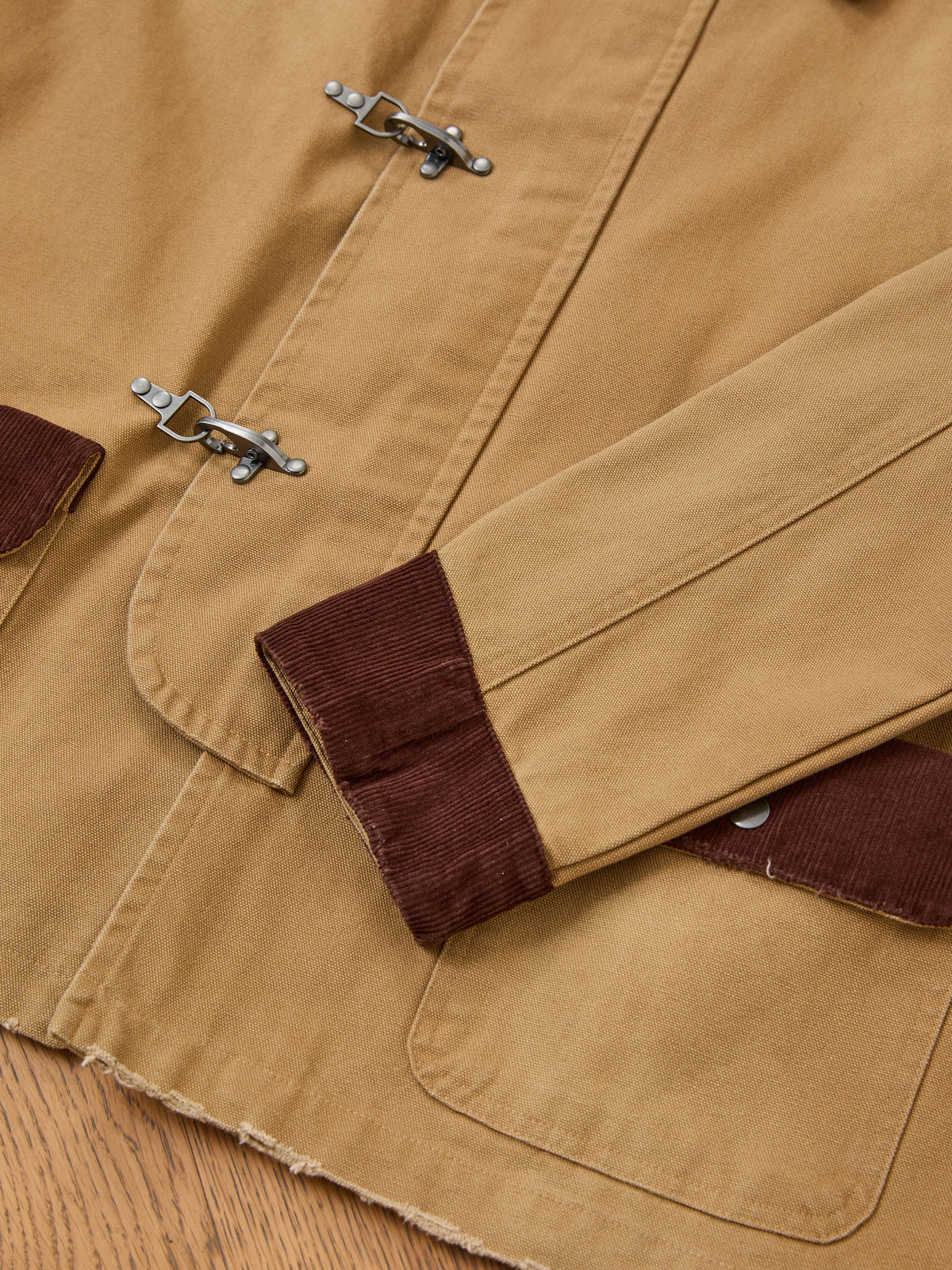 The Fireman Clasp Canvas Work Jacket by Found is a unisex, relaxed fit, mid-weight everyday staple crafted from tan washed canvas with brown corduroy cuffs and pocket flaps. Featuring signature metal toggle clasps on the front, this jacket is displayed laid out on a wooden surface.