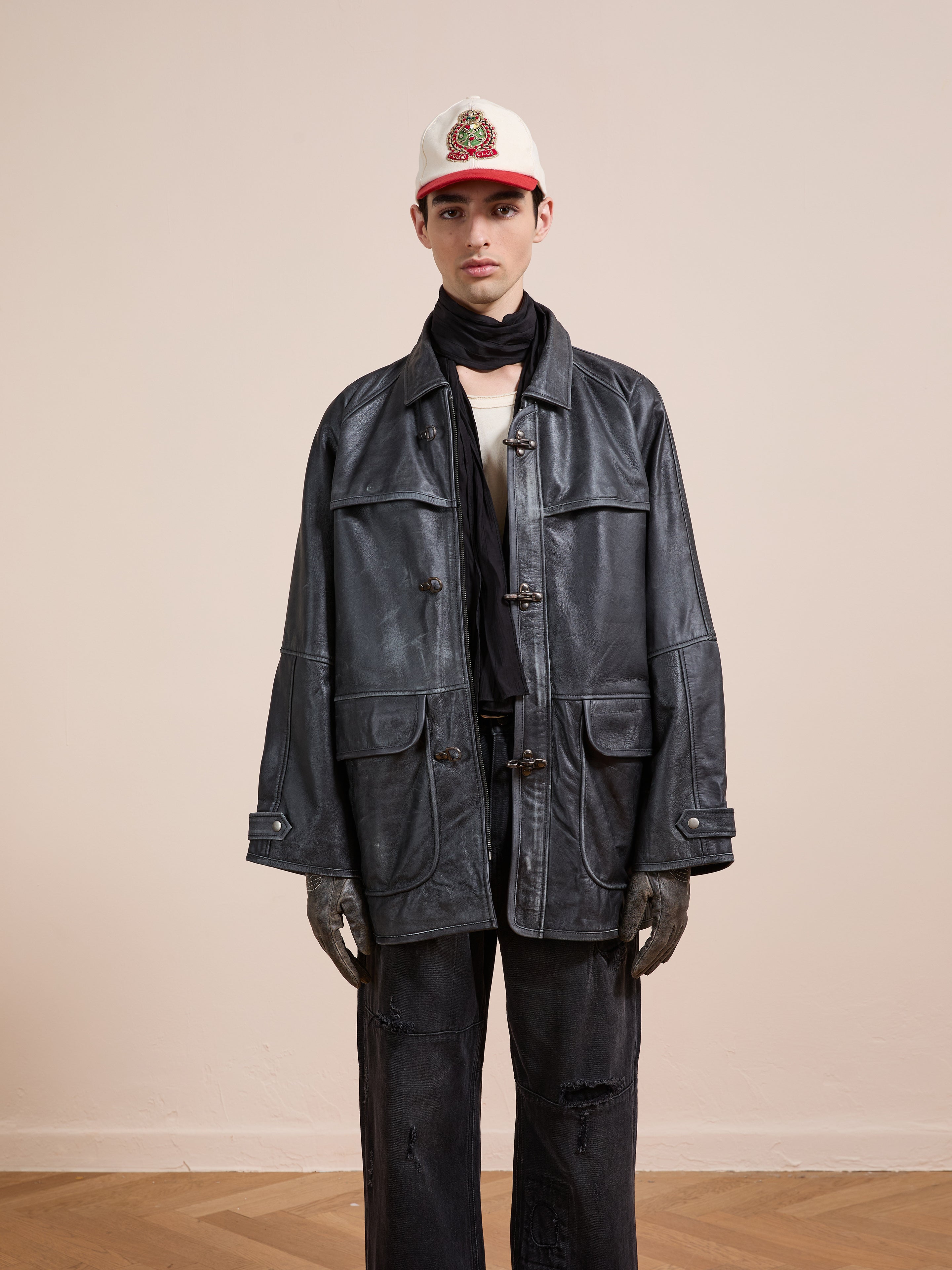 A person wearing the FOUND Distressed Leather Fireman Clasp Jacket stands against a plain beige wall, styled with a black scarf, white emblem cap, black gloves, and distressed black pants.