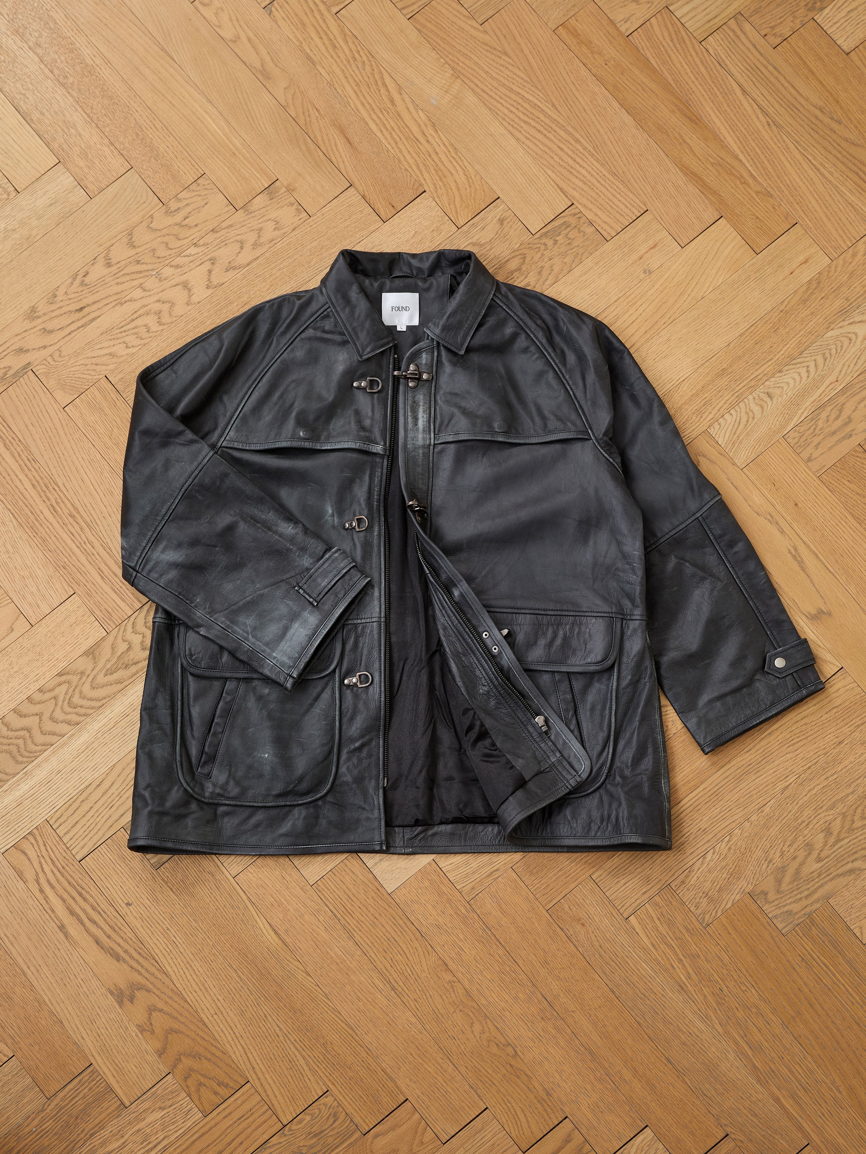 The FOUND Distressed Leather Fireman Clasp Jacket is laid flat on a light wooden herringbone floor.
