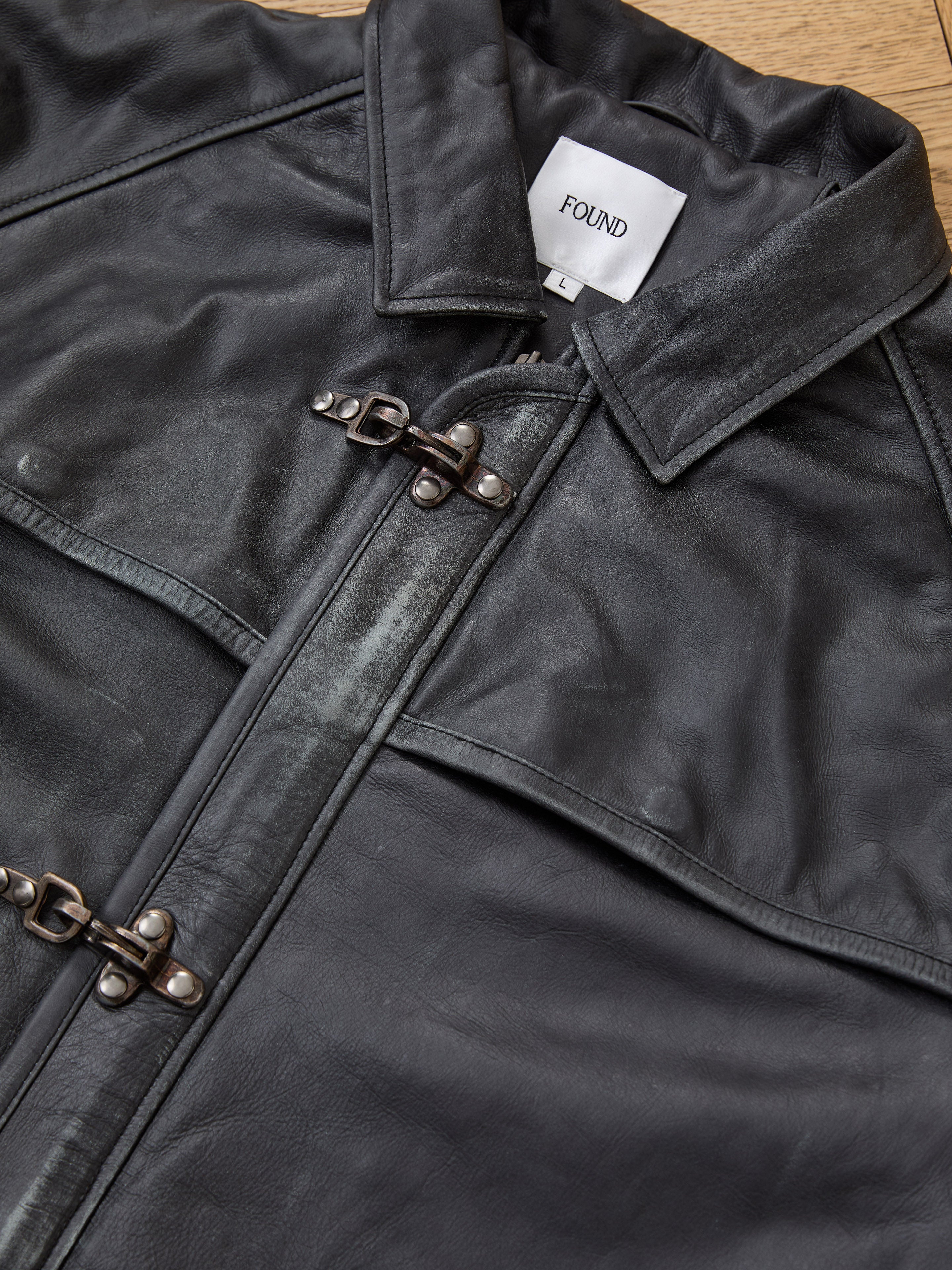 Close-up of the FOUND Distressed Leather Fireman Clasp Jacket, a unisex design made from genuine black leather, featuring a collar, two clasp closures, and a white "FOUND" label inside the neckline for timeless style.