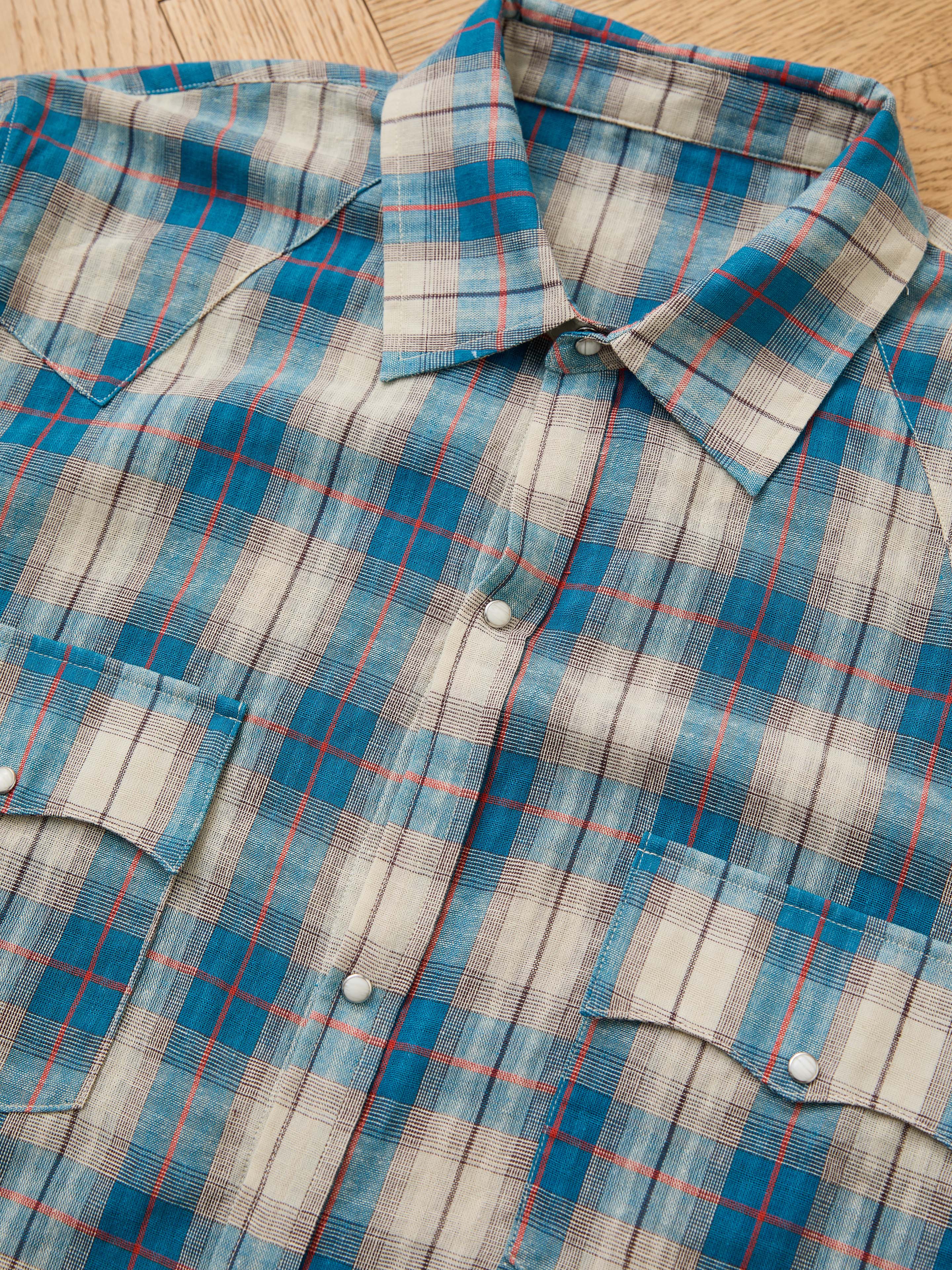 The FOUND Flannel Shirt, in a blue, white, and red Western plaid with two front pockets and a classic unisex relaxed fit, is laid flat on a wooden surface.