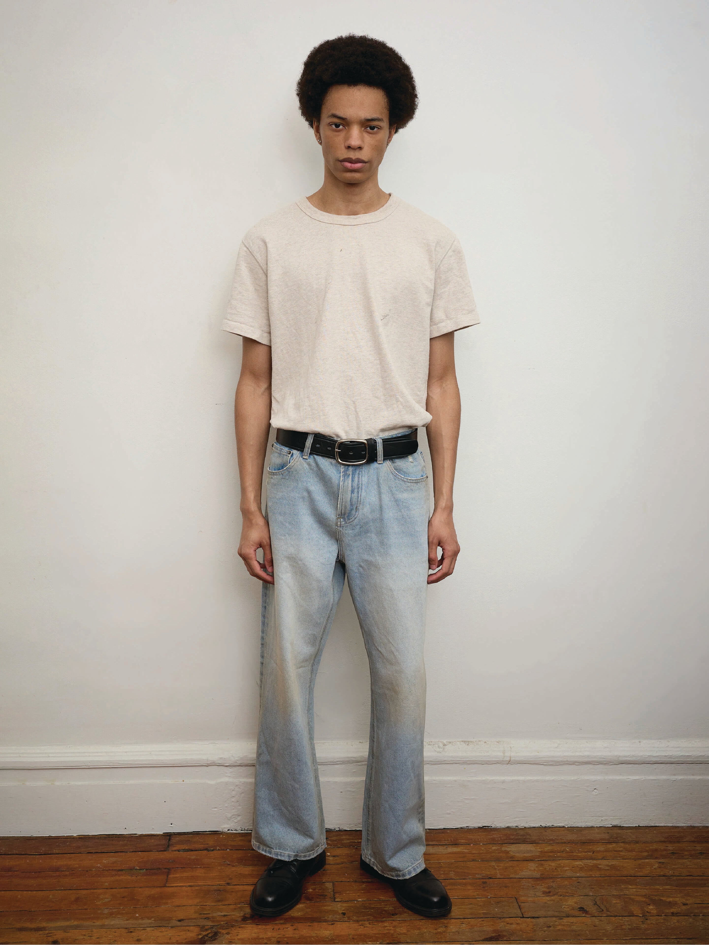 A person stands against a plain white wall wearing a beige t-shirt, Found Flare Jeans in light blue denim, a black belt, and black shoes on a wooden floor.