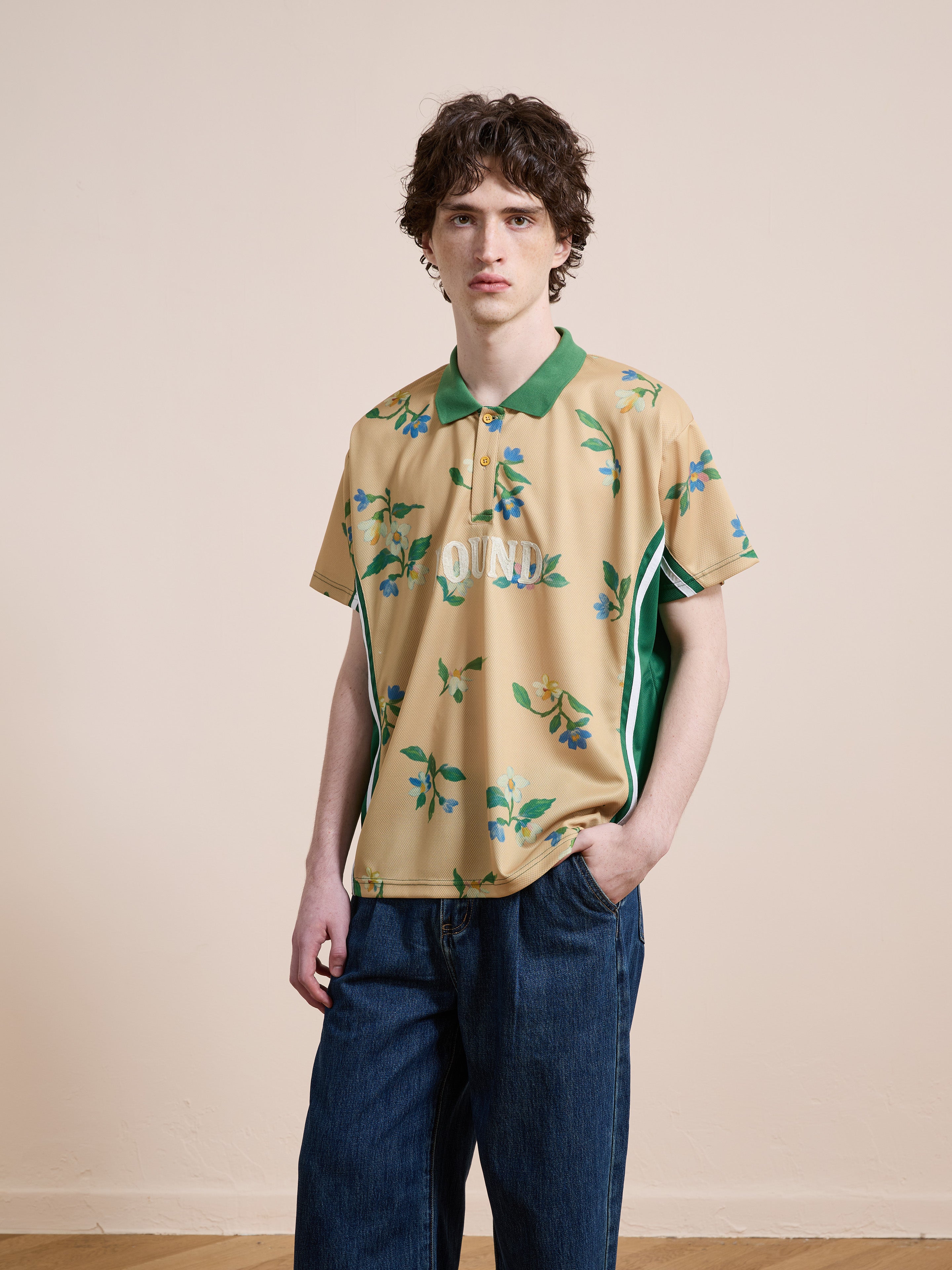 A person with curly brown hair stands against a plain background, wearing the FOUND Floral Polo Jersey—a vintage-style unisex polo with green accents—paired with relaxed fit blue jeans, one hand tucked in their pocket.
