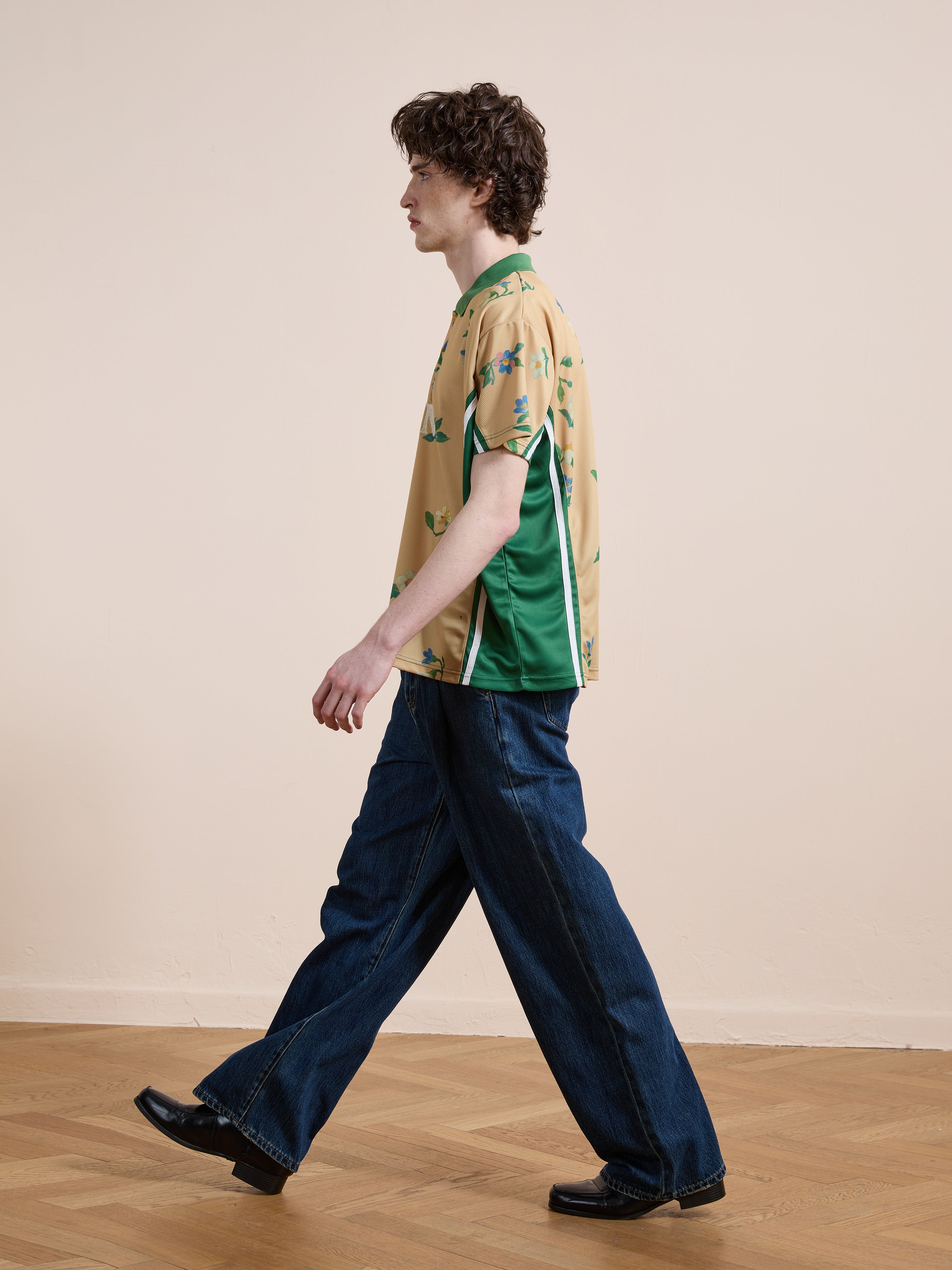 A person with curly hair walks indoors on a wooden floor wearing the FOUND Floral Polo Jersey, wide-leg blue jeans, and black shoes.