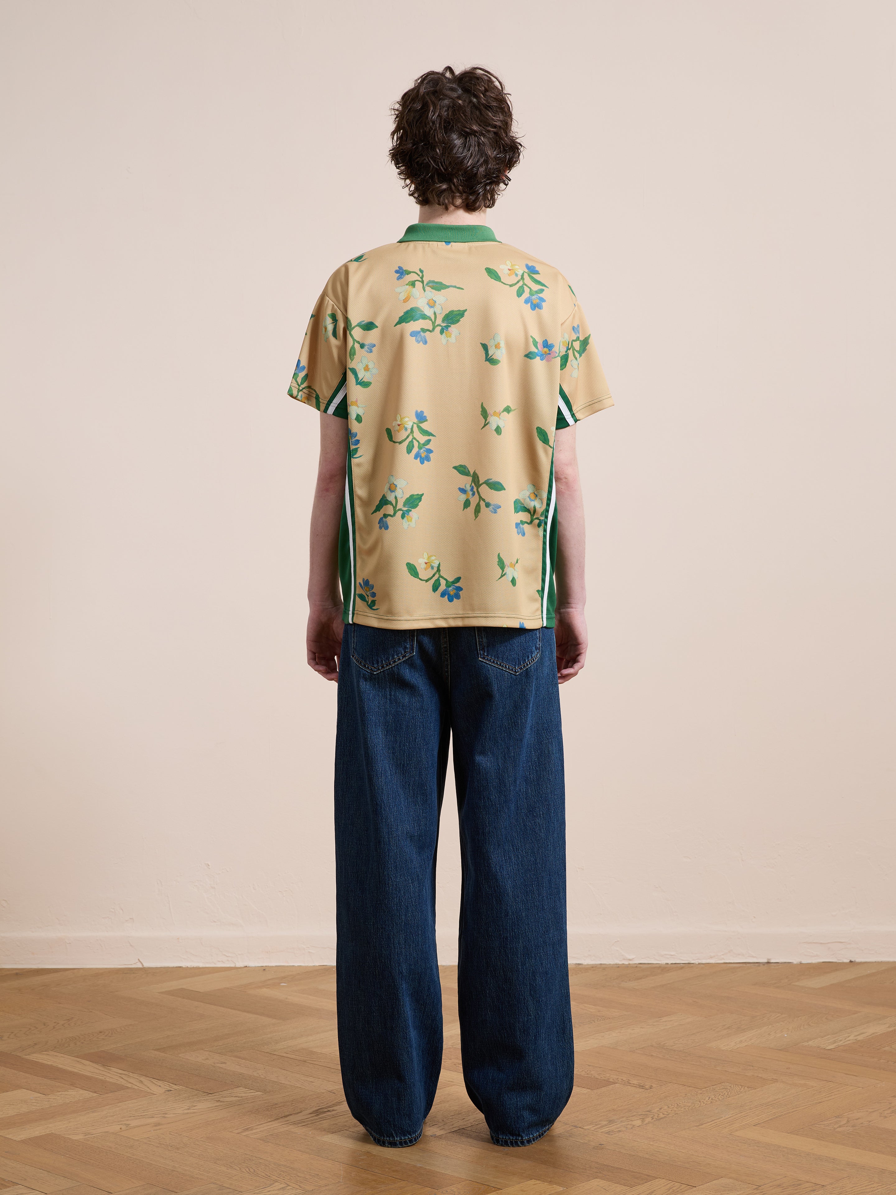 A person stands indoors on a wooden floor, facing away, wearing the FOUND Floral Polo Jersey with green prints in a unisex relaxed fit, paired with loose-fitting blue jeans.