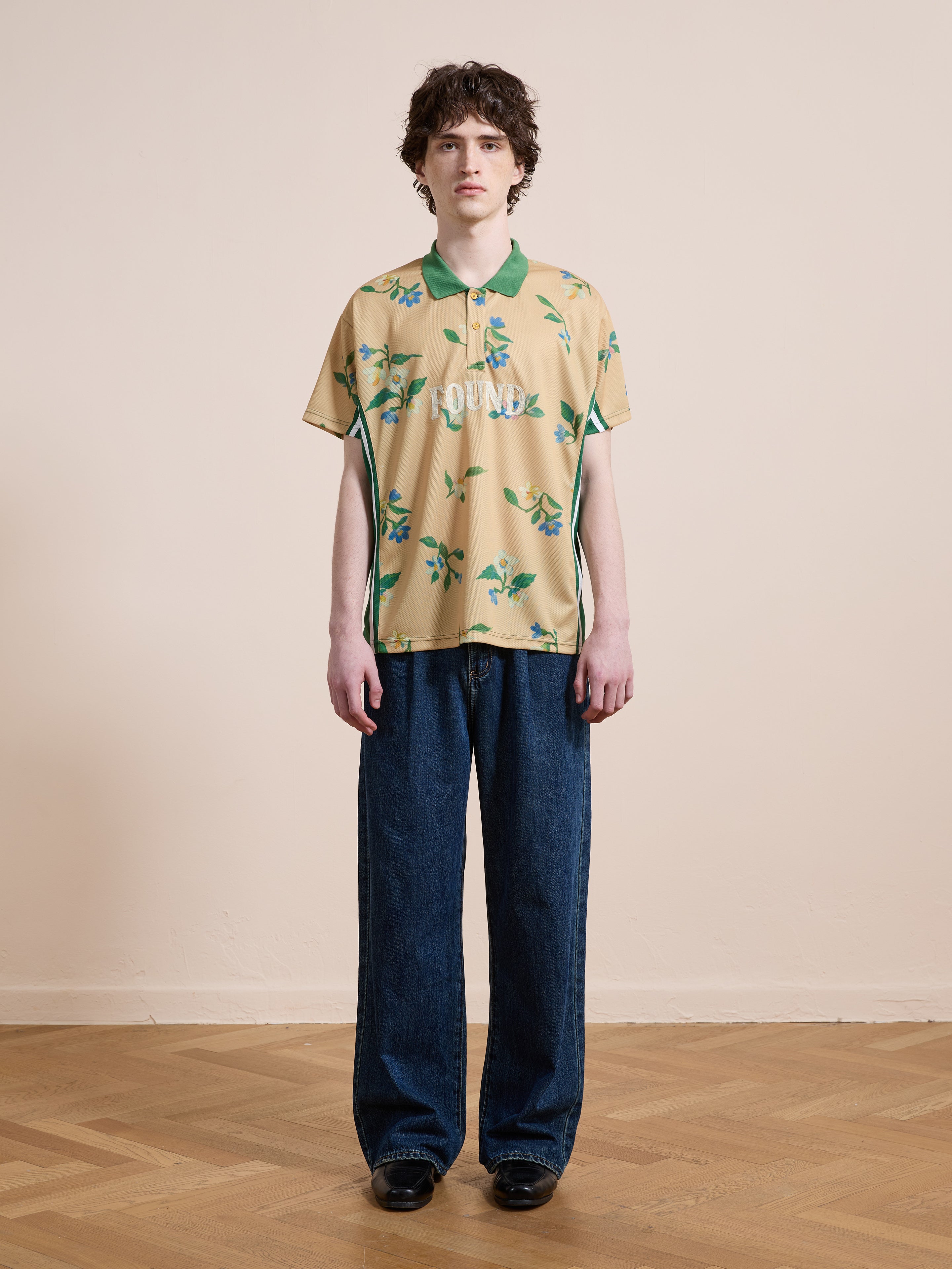 Indoors, a person wears the FOUND Floral Polo Jersey in yellow, paired with wide blue jeans and black shoes, standing against a beige wall and wooden floor.