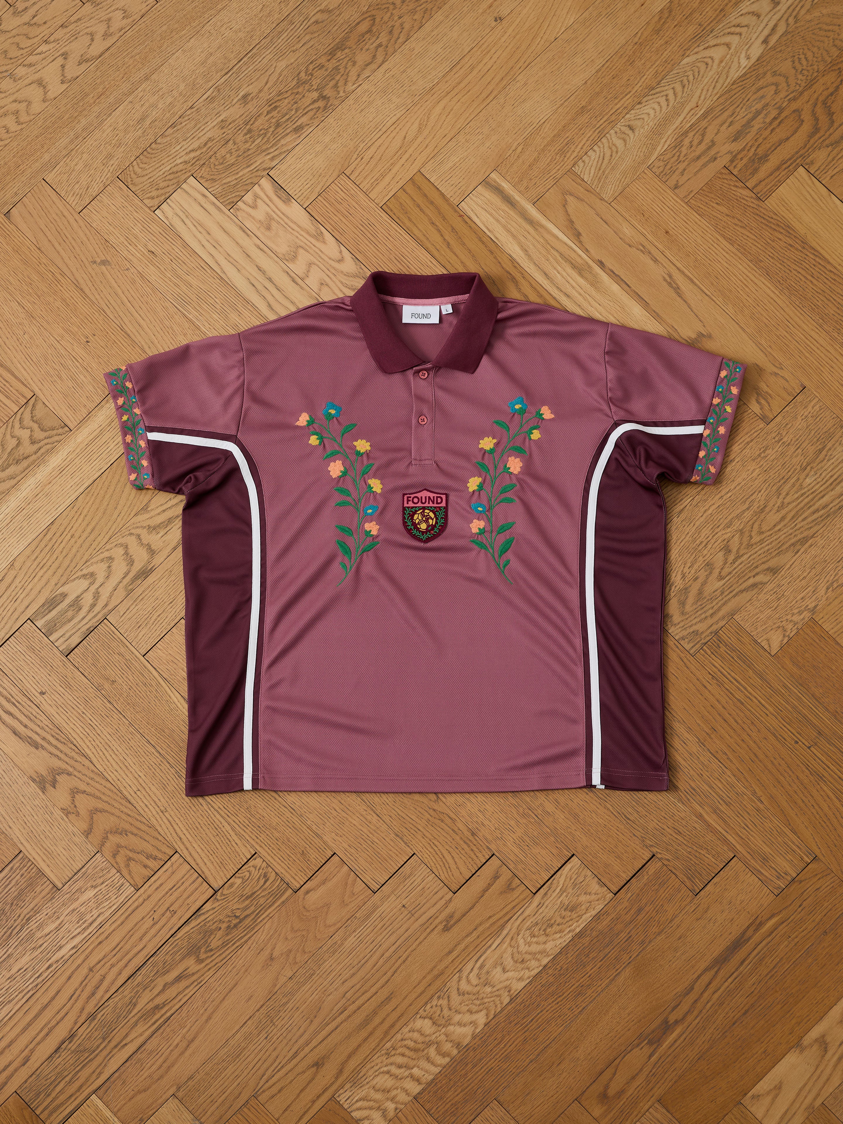 The FOUND Floral Polo Jersey, a maroon cotton polo with floral embroidery on the front and sleeves and a "FOUND" patch, is displayed on a patterned wooden floor.